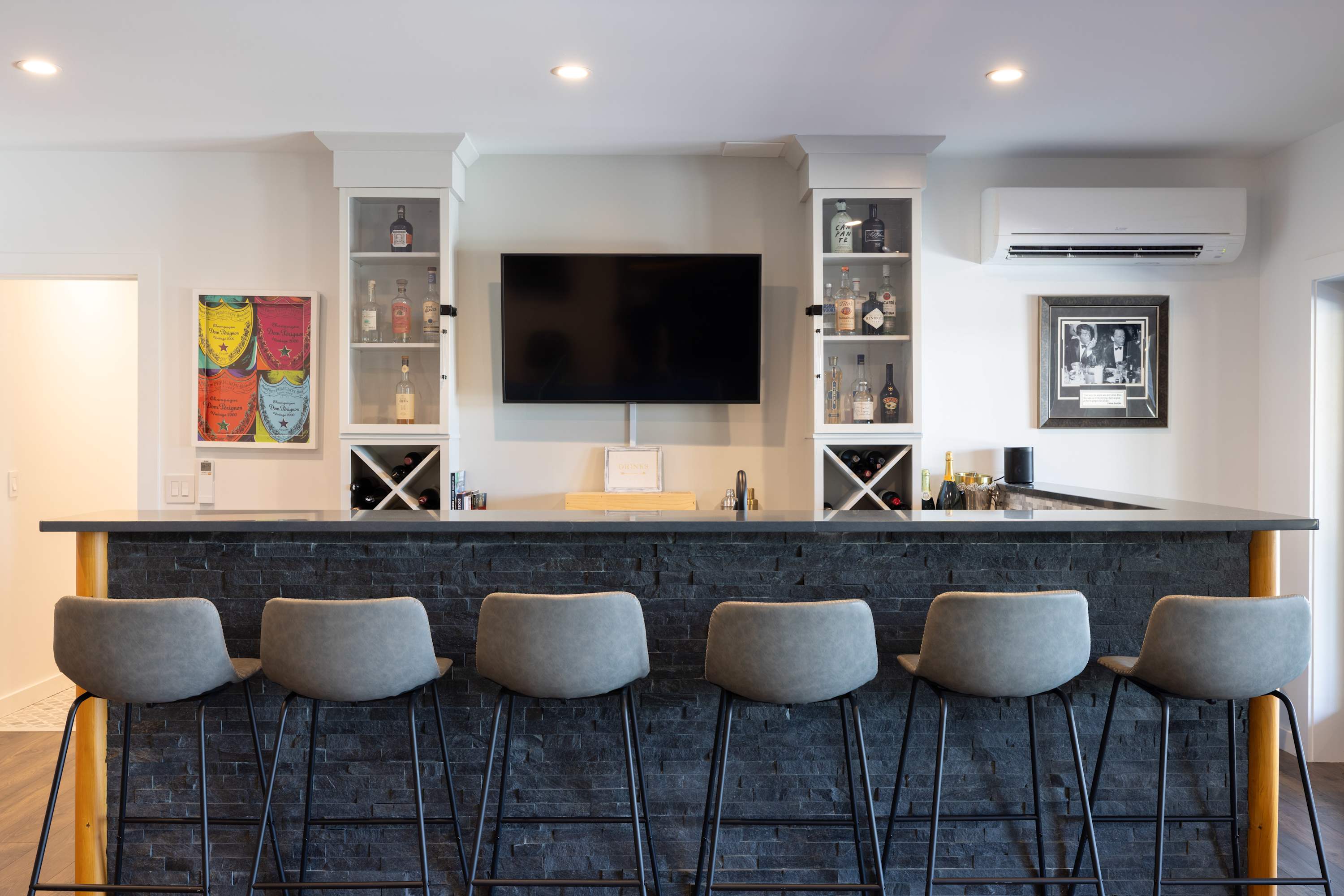 Modern wet bar with a dark stone counter and stacked stone facade, featuring six grey bar stools, built-in display cabinets with liquor bottles, a mounted TV, and recessed lighting.