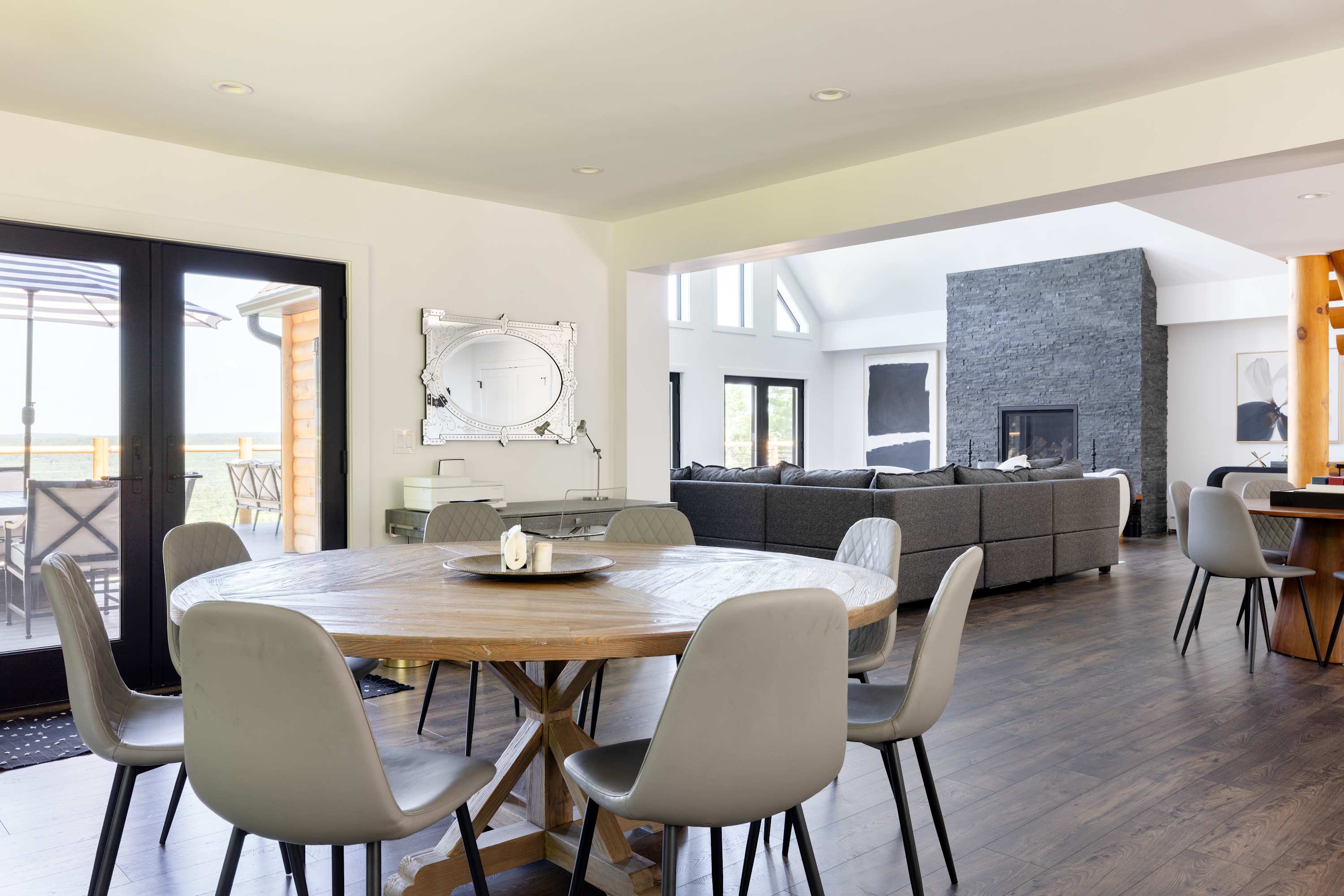 A spacious dining area with a round wooden table and six gray upholstered chairs, featuring dark hardwood floors and an open view into the living room with a stone fireplace.