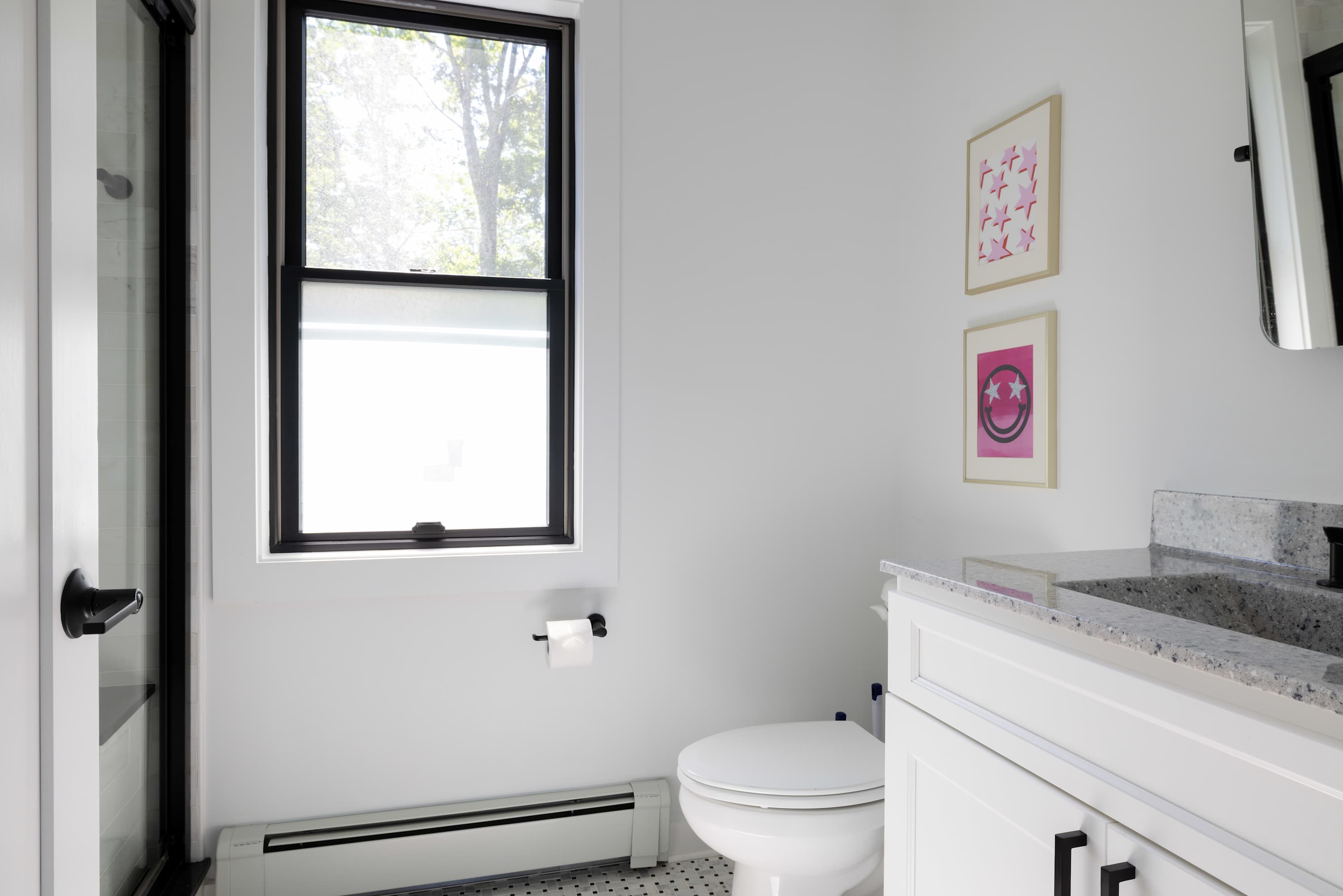 Bright bathroom with white walls, black and white tiled floor, and a white vanity with a gray speckled countertop. A toilet is next to a window with black trim, and colorful pop art hangs on the wall.