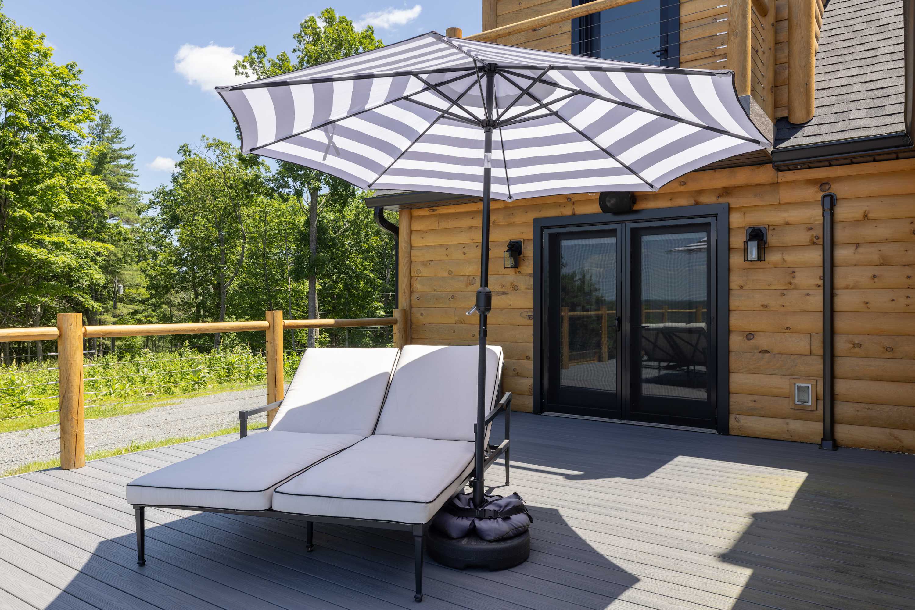 A gray composite deck features two white lounge chairs with black piping under a large striped umbrella, positioned near the wooden-sided house with black French doors, surrounded by lush trees.