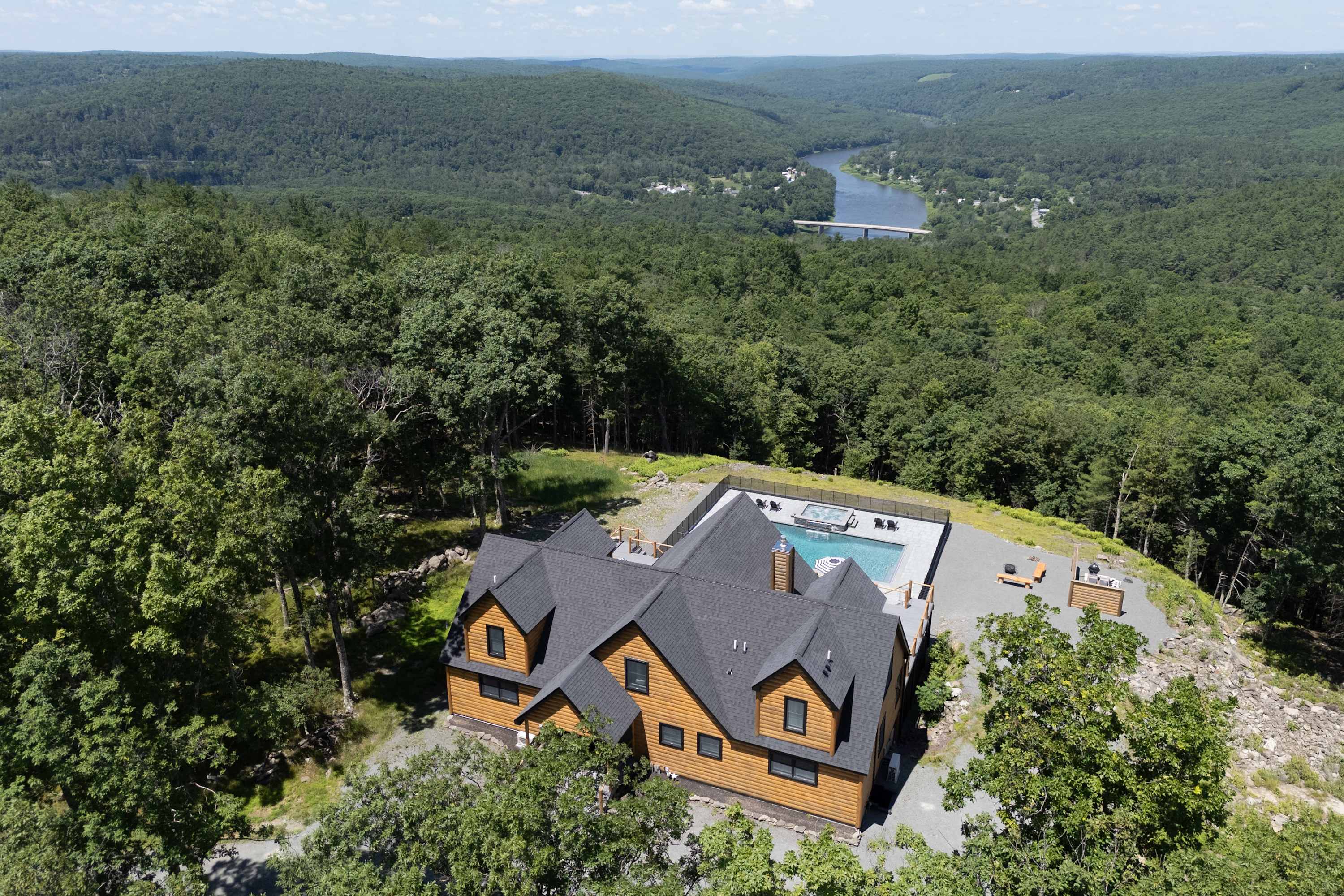 An aerial view of a large log cabin-style house with a dark gray roof, nestled amidst a dense green forest. A rectangular swimming pool and hot tub area are visible on a patio next to the house, overlooking the expansive landscape.