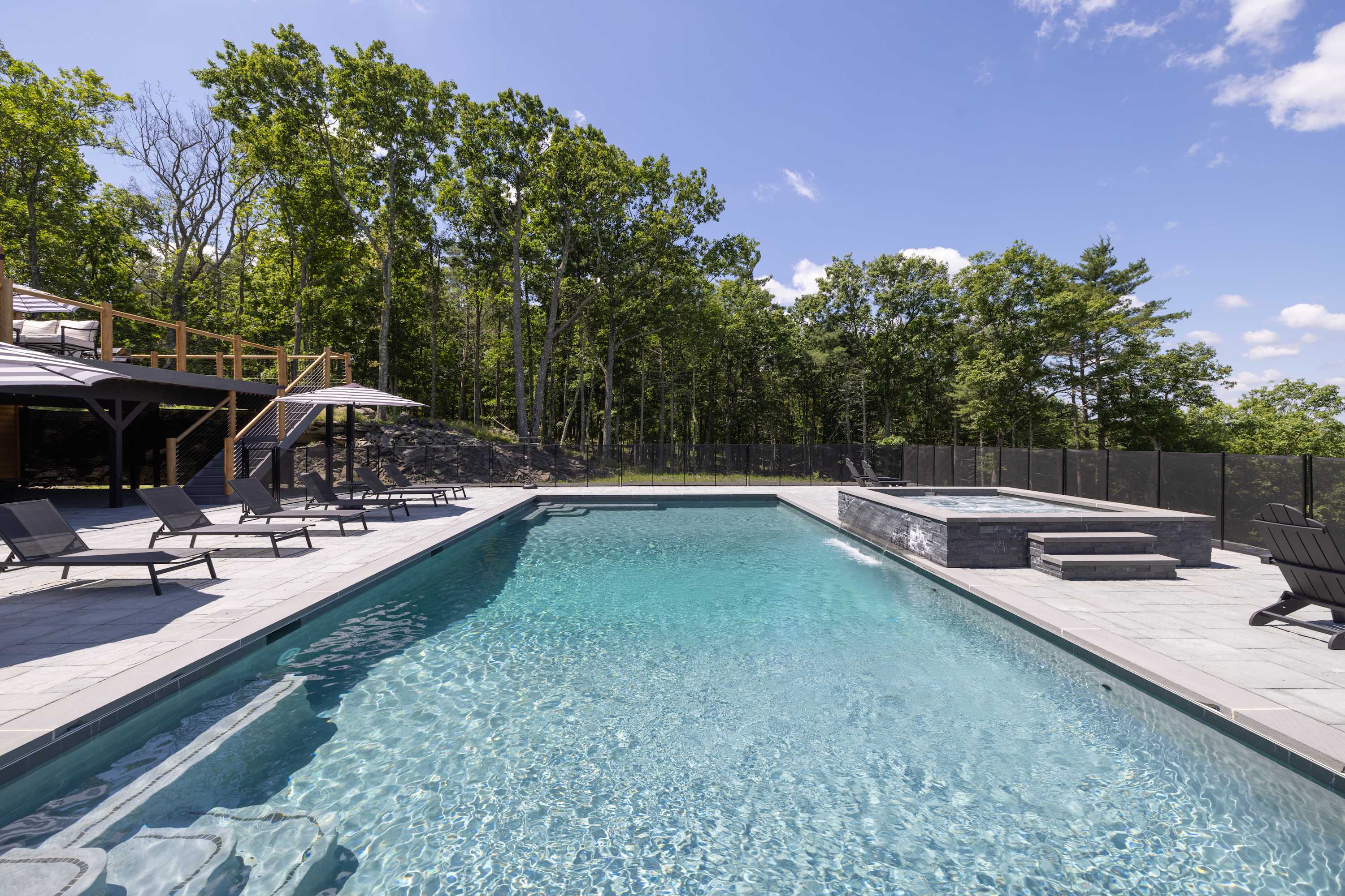 Expansive outdoor swimming pool with an integrated hot tub featuring a waterfall, surrounded by a paved patio with lounge chairs and lush green trees under a bright blue sky.