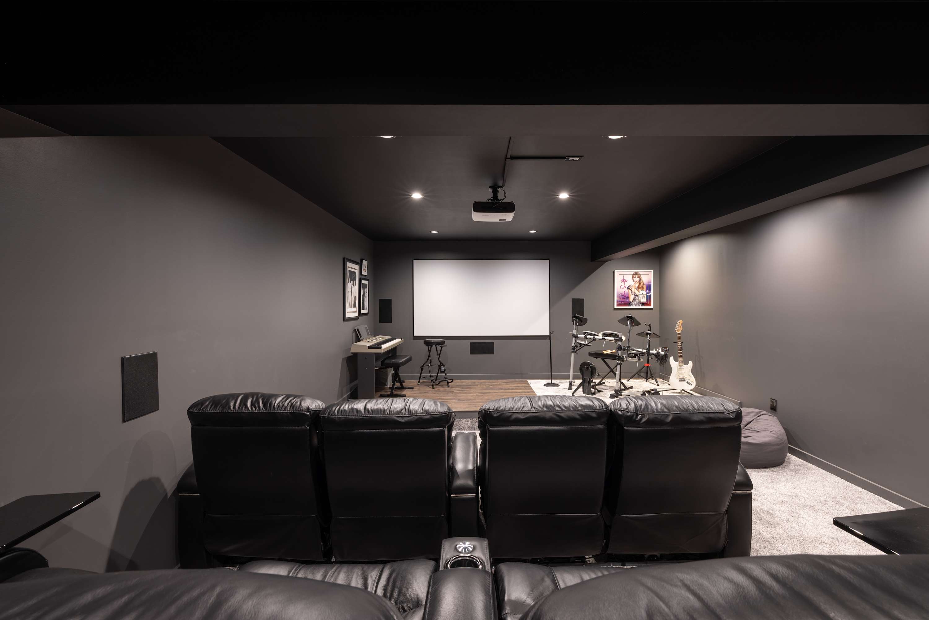 Dark-themed media room featuring a large projection screen, comfortable black leather recliners, and a stage area with musical instruments like a keyboard, drum set, and guitars.