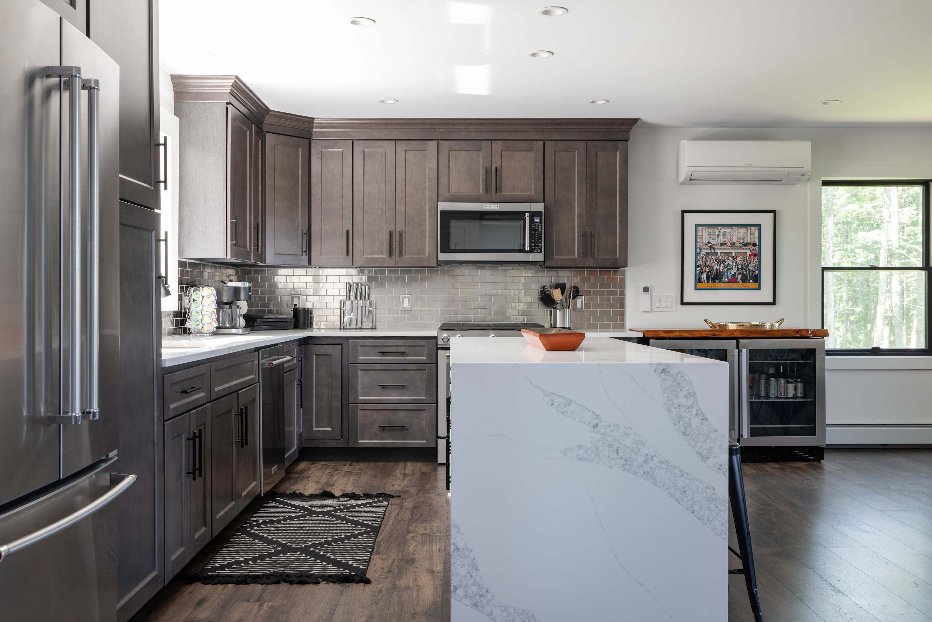 A modern kitchen with dark gray cabinets, stainless steel appliances, a white quartz waterfall island, and a metallic subway tile backsplash, with a mini-split AC unit visible above a window.