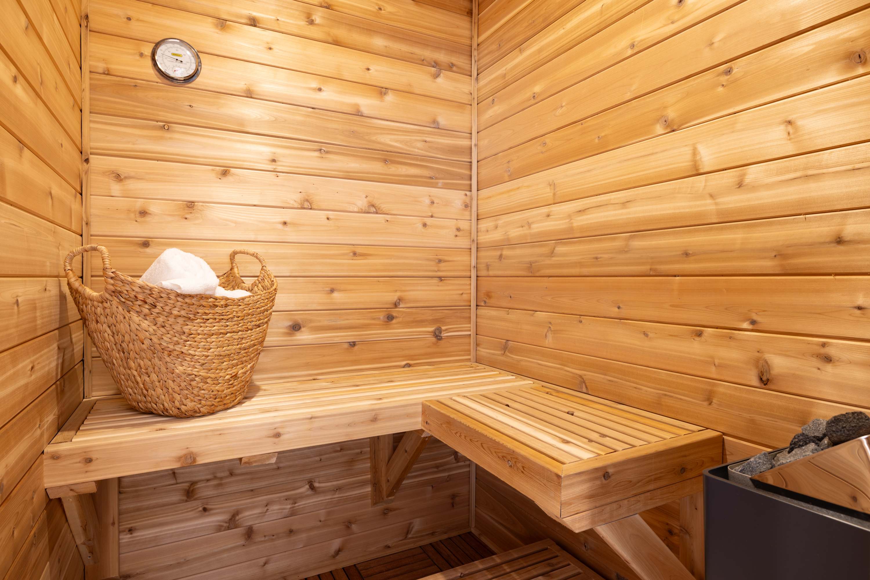 Cozy indoor sauna with light cedar wood paneling on walls and benches, featuring a thermometer and a woven basket with towels, creating a warm and inviting atmosphere.