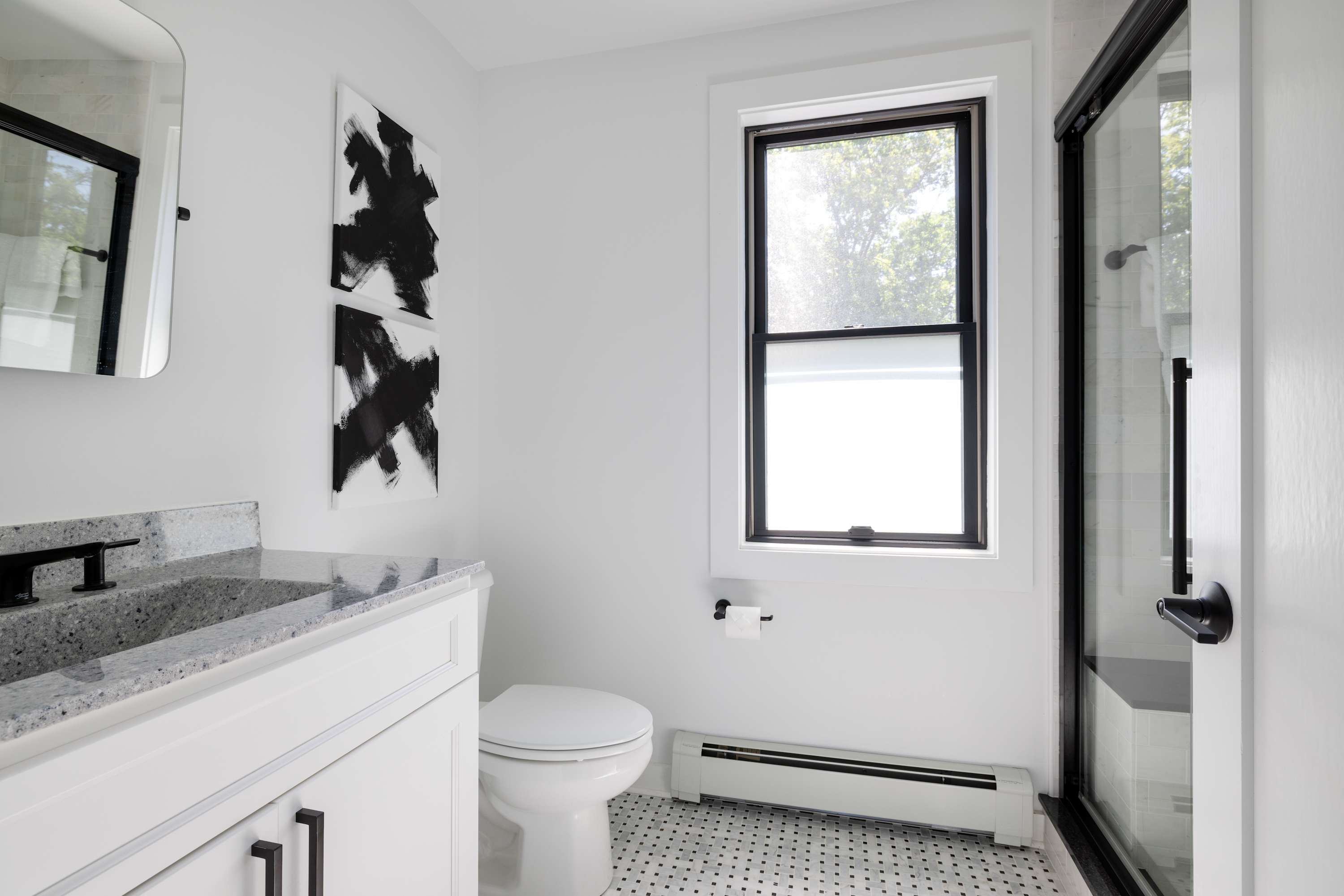Modern bathroom with white walls, black and white tiled floor, and a white vanity with a gray speckled countertop. A toilet is next to a window with black trim, and a glass shower enclosure is visible.