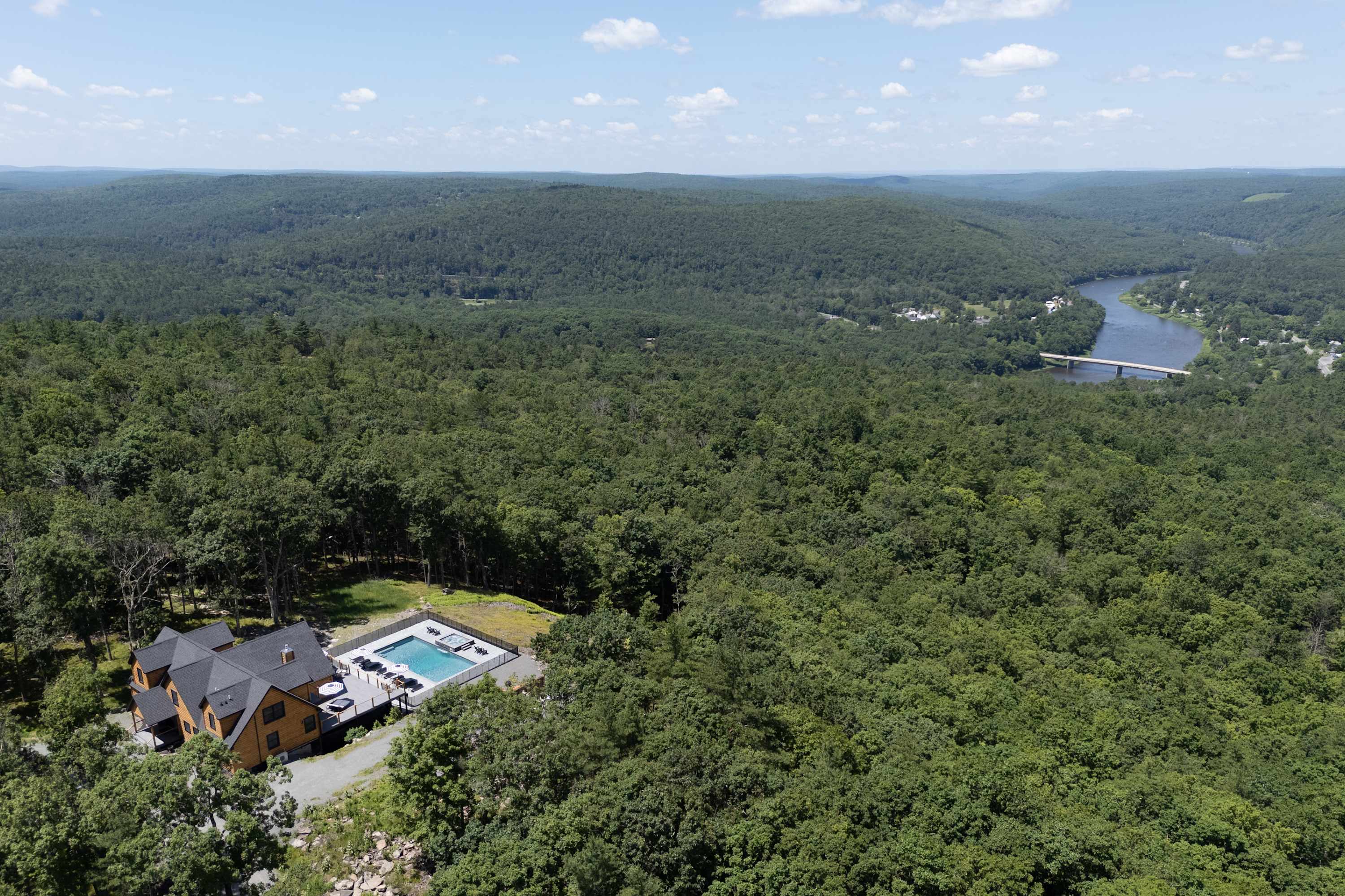 A high aerial view captures the log cabin-style house with its dark roof, surrounded by a vast expanse of lush green forest. The outdoor pool and hot tub area are clearly visible on the hilltop property, overlooking a distant river and valley.