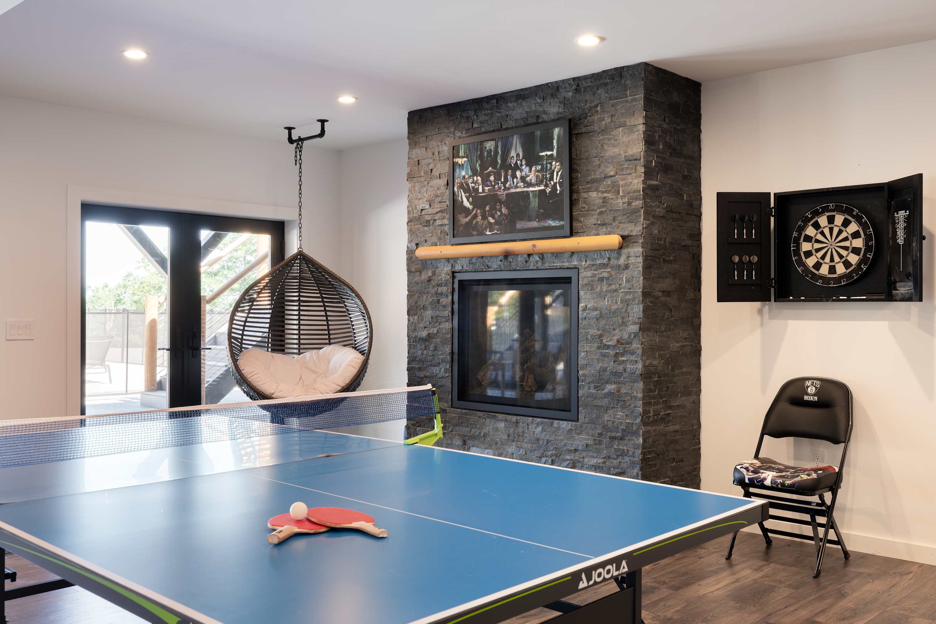 Bright game room featuring a blue ping pong table, a modern stone fireplace with a mounted TV, and a hanging egg chair, offering various entertainment options.