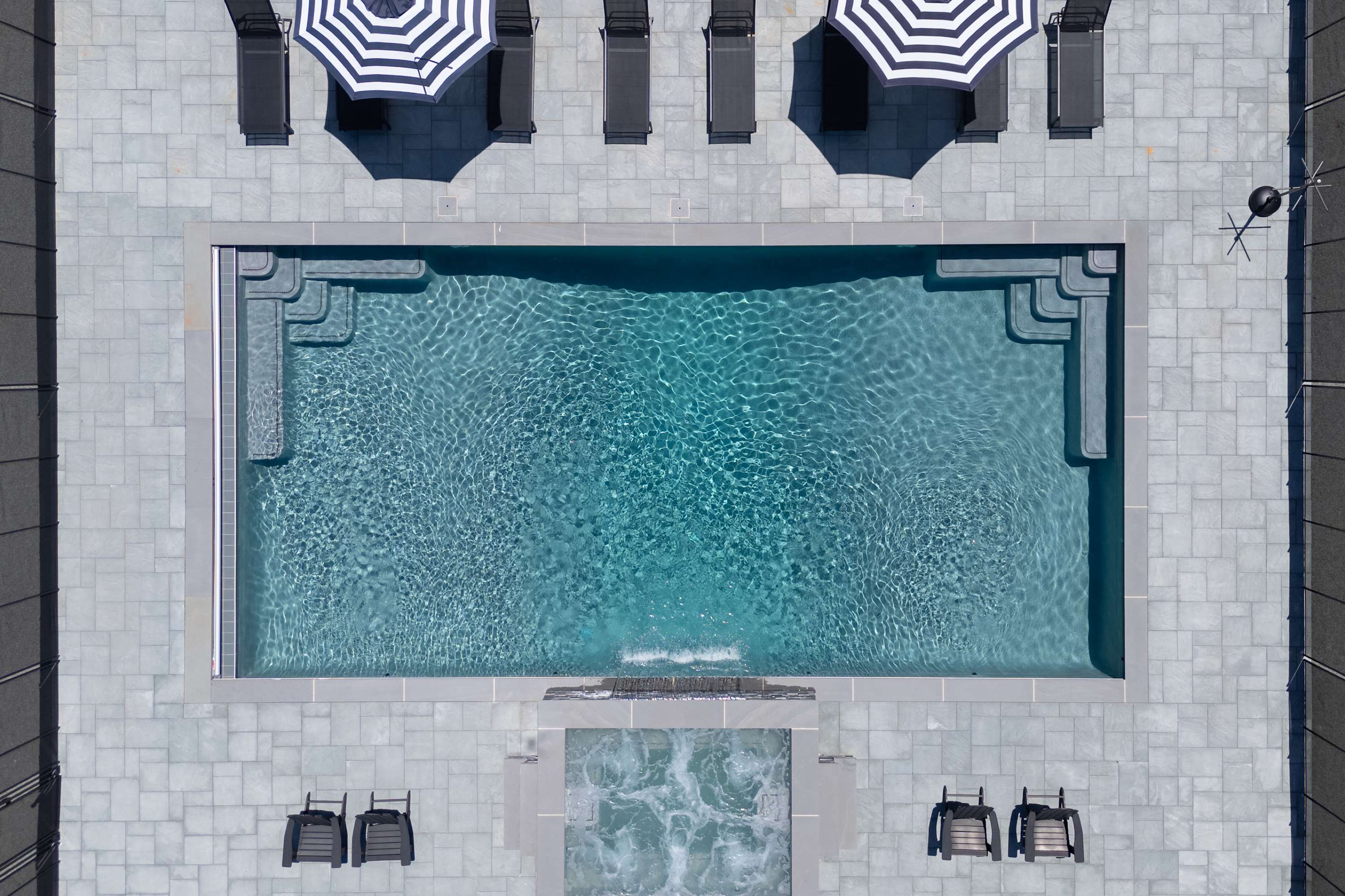 An aerial view of a rectangular swimming pool with light blue water and integrated steps, surrounded by gray paver stones. A separate hot tub with bubbling water is adjacent, with lounge chairs and striped umbrellas nearby.