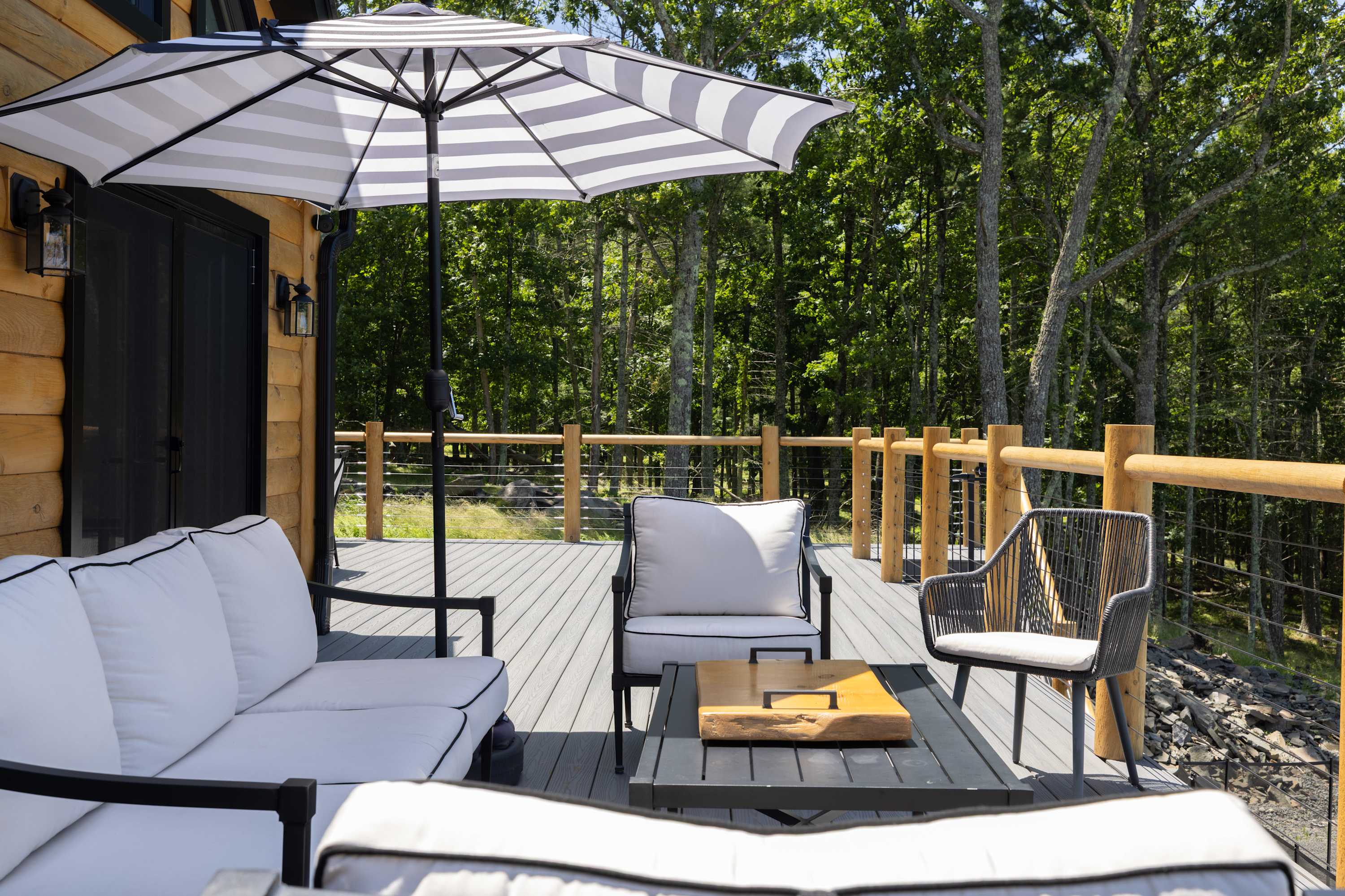 A spacious outdoor deck with gray composite flooring and a wooden railing, furnished with a striped umbrella, white outdoor sofa, and chairs, offering a serene view of a dense forest.