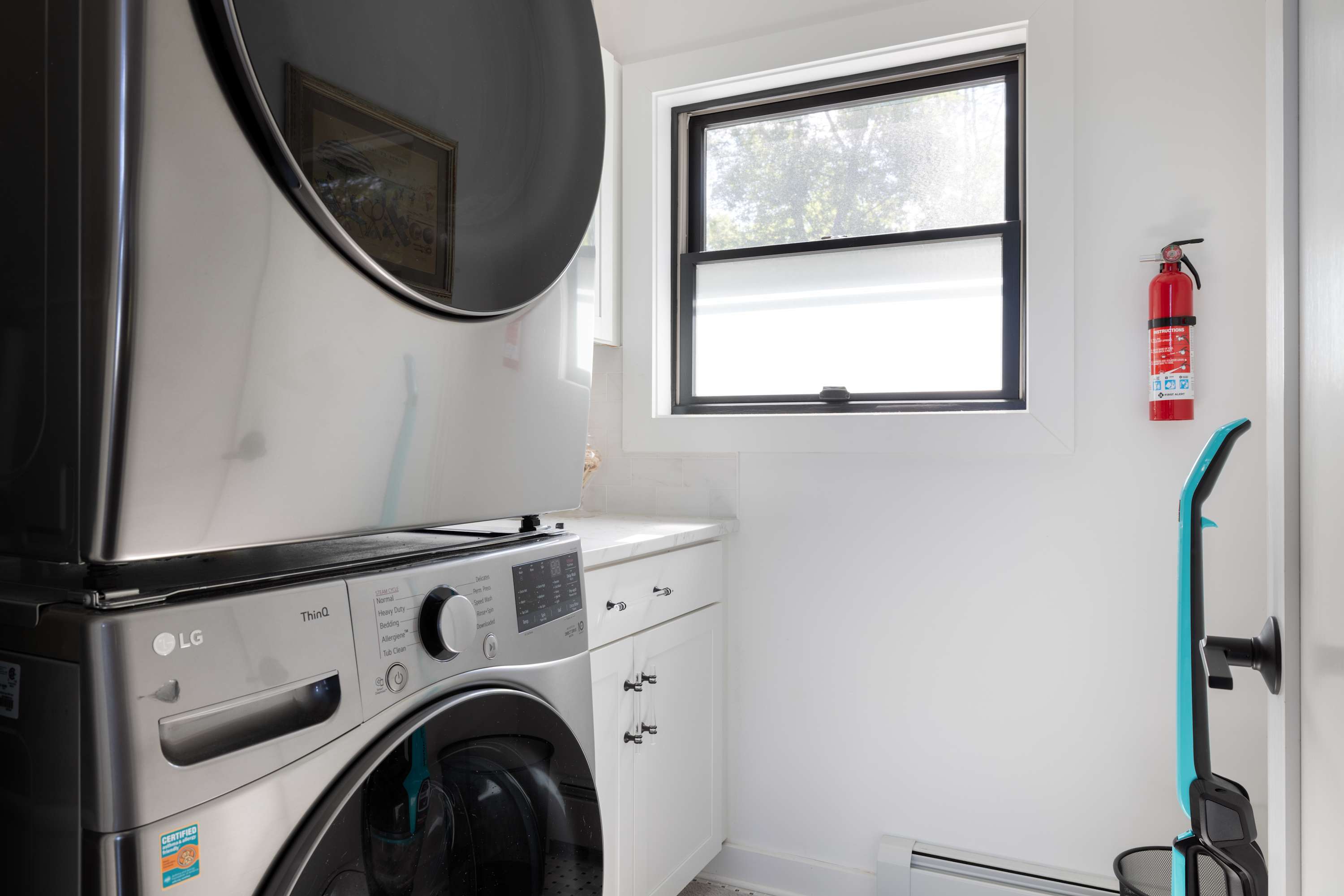 Bright laundry room with stacked silver washer and dryer units, white cabinets, a light countertop, and a window, featuring a fire extinguisher mounted on the wall.
