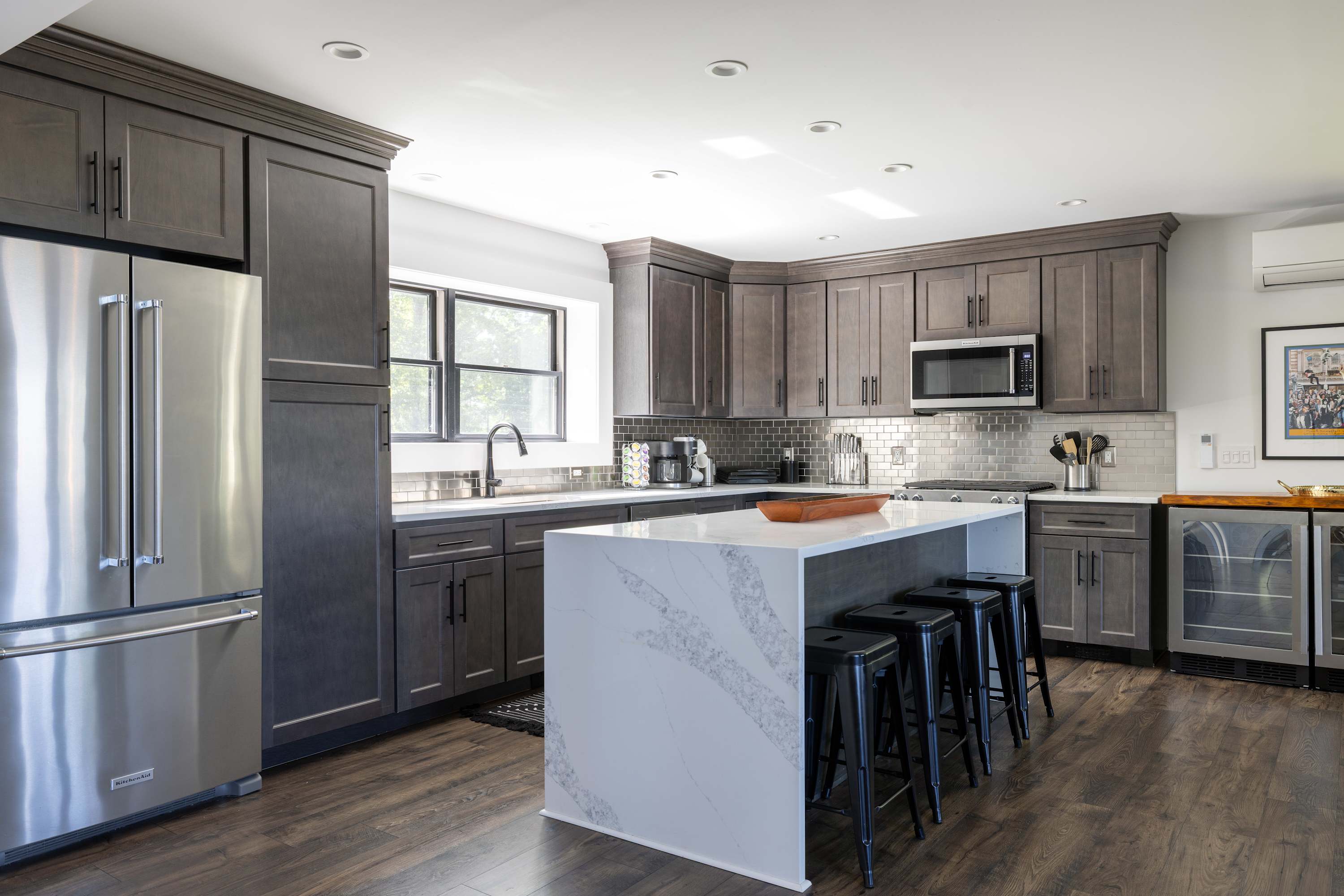 A contemporary kitchen featuring dark gray shaker cabinets, stainless steel appliances, a white quartz waterfall island with black bar stools, and a metallic subway tile backsplash.