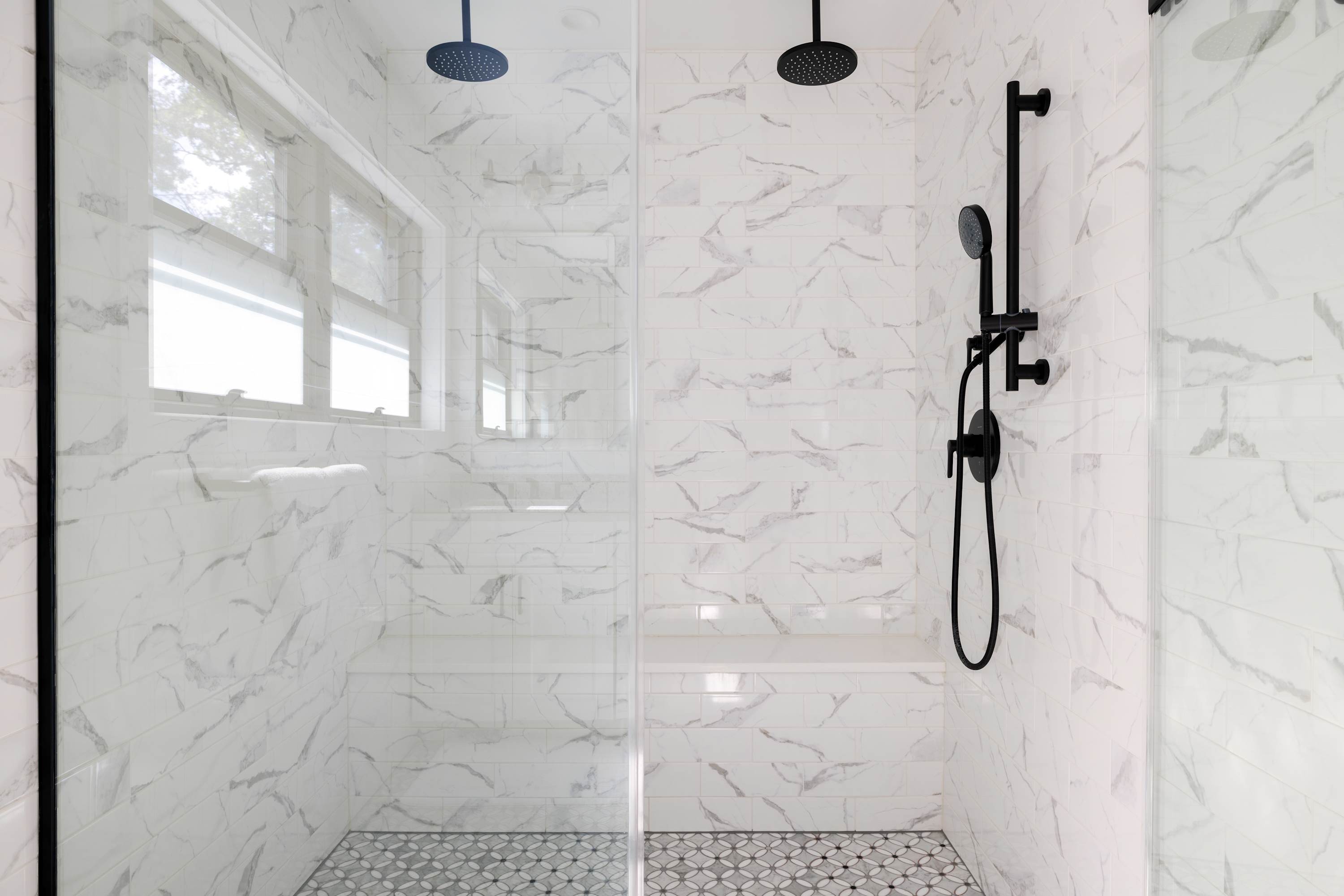 A spacious walk-in shower with white marble-look subway tiles, black rain shower heads, and a built-in bench, featuring a patterned floor tile and clear glass enclosure.