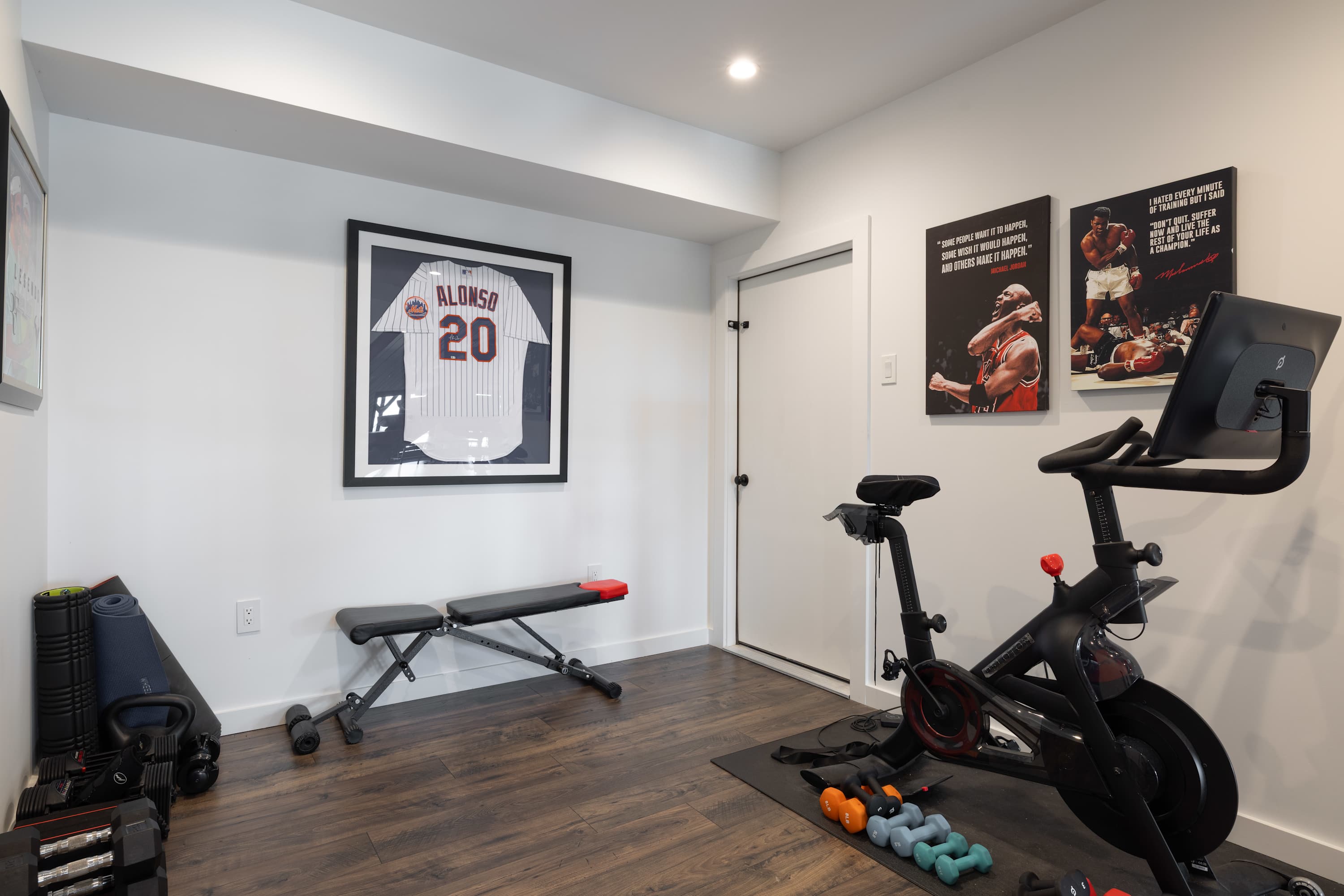 Home gym with dark wood-look flooring, featuring a Peloton bike, a weight bench, dumbbells, and framed sports memorabilia on white walls.
