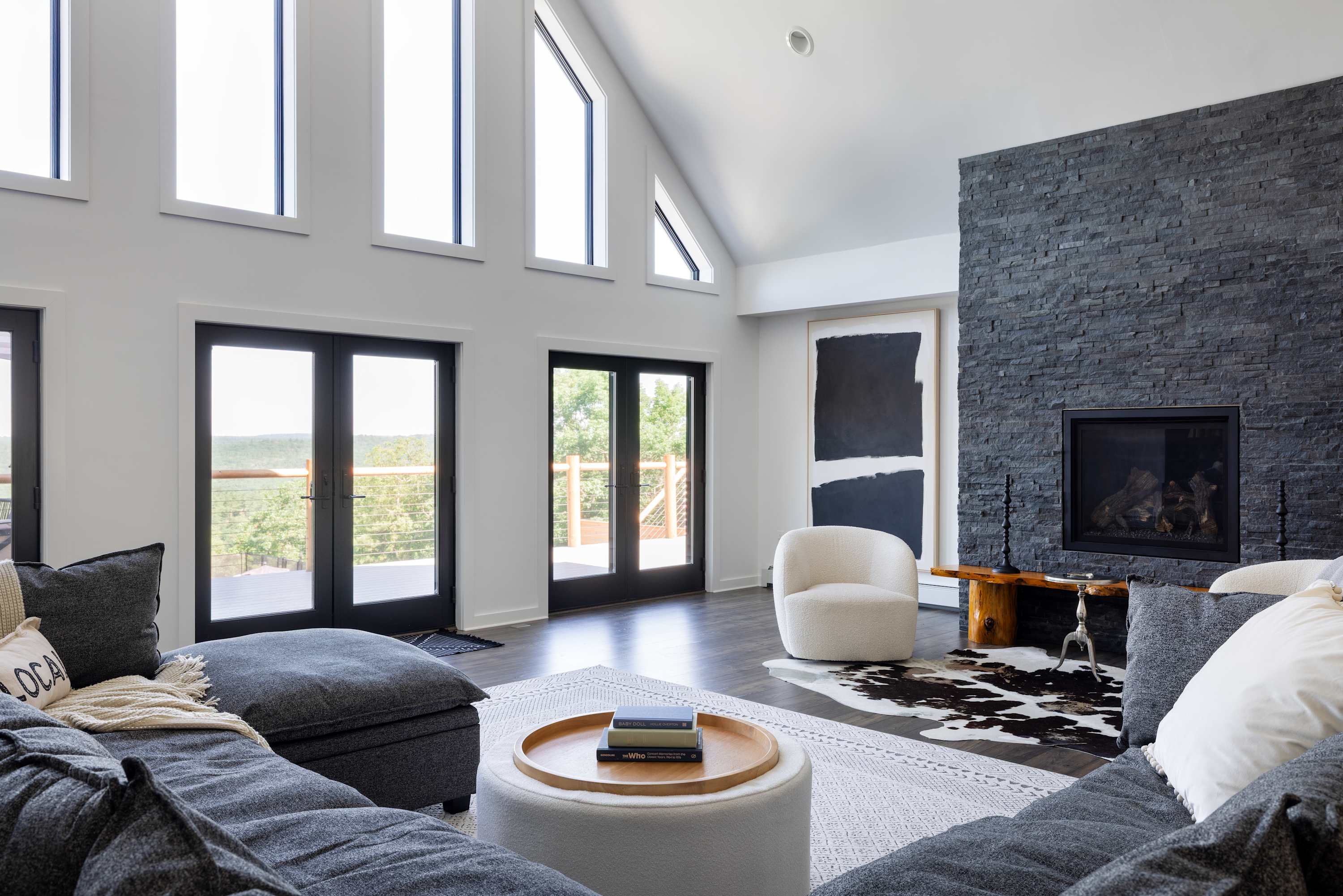 A modern living room with a large gray sectional sofa, a white ottoman coffee table, and a dark stone fireplace, featuring a cowhide rug and large windows overlooking a wooded area.