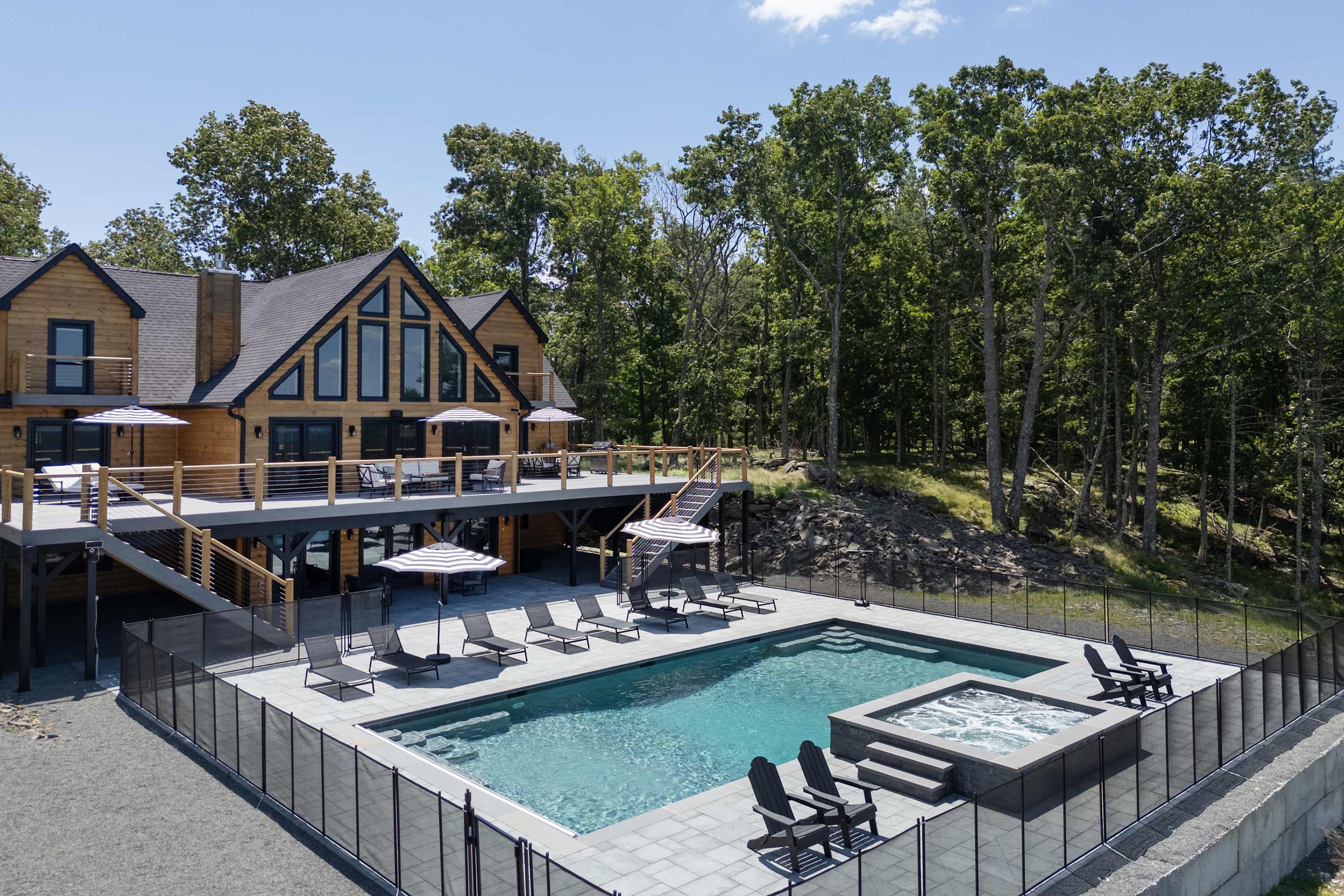 Aerial view of a modern wooden lodge with extensive decking, a large rectangular swimming pool, and an adjacent hot tub, set amidst a dense forest under a bright sunny sky.