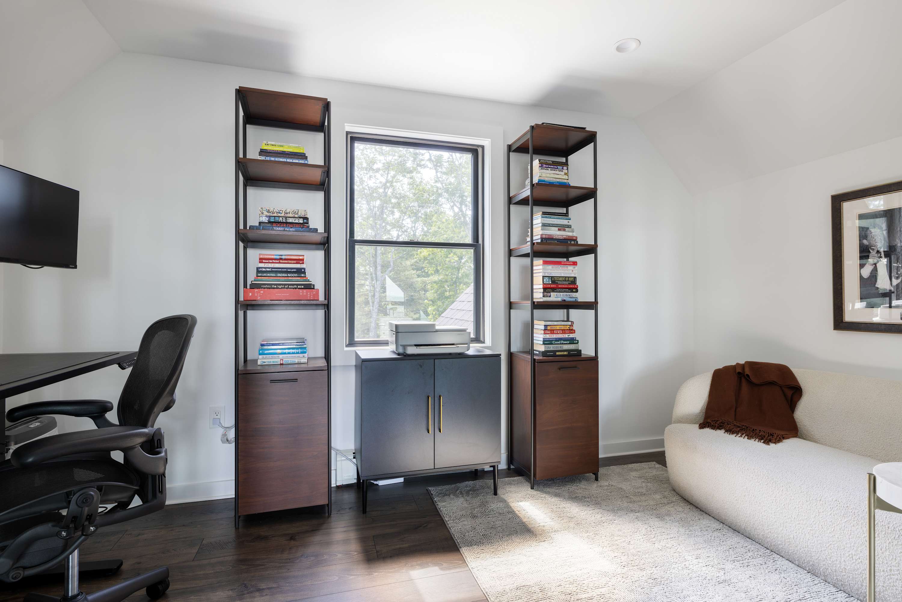 Home office with dark wood floors, two tall dark wood and metal bookshelves flanking a window, and a dark cabinet below the window. A black office chair and desk are on the left, and a white textured sofa is on the right.