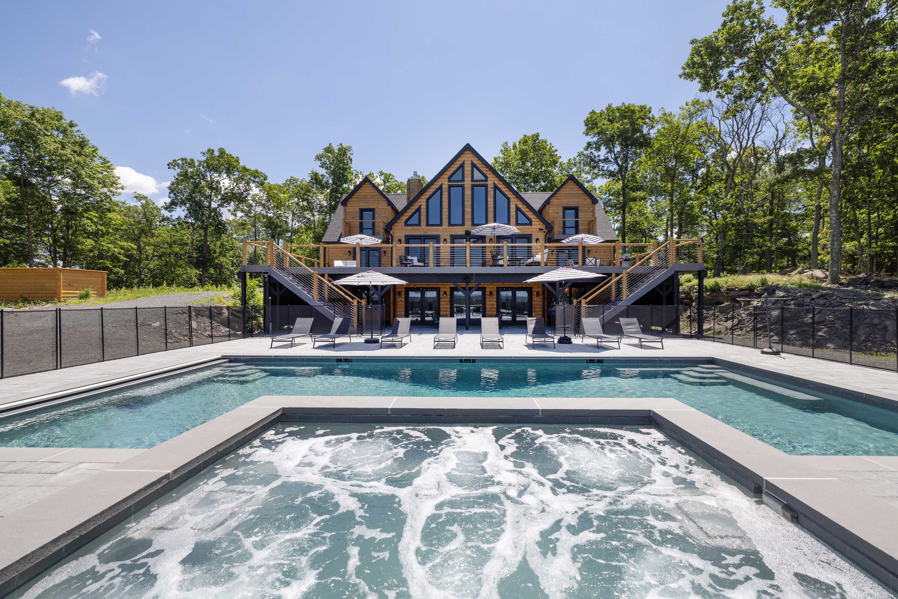 Grand wooden lodge-style house with large windows and multiple decks, overlooking a spacious pool area with a built-in hot tub, surrounded by lush green trees under a clear blue sky.