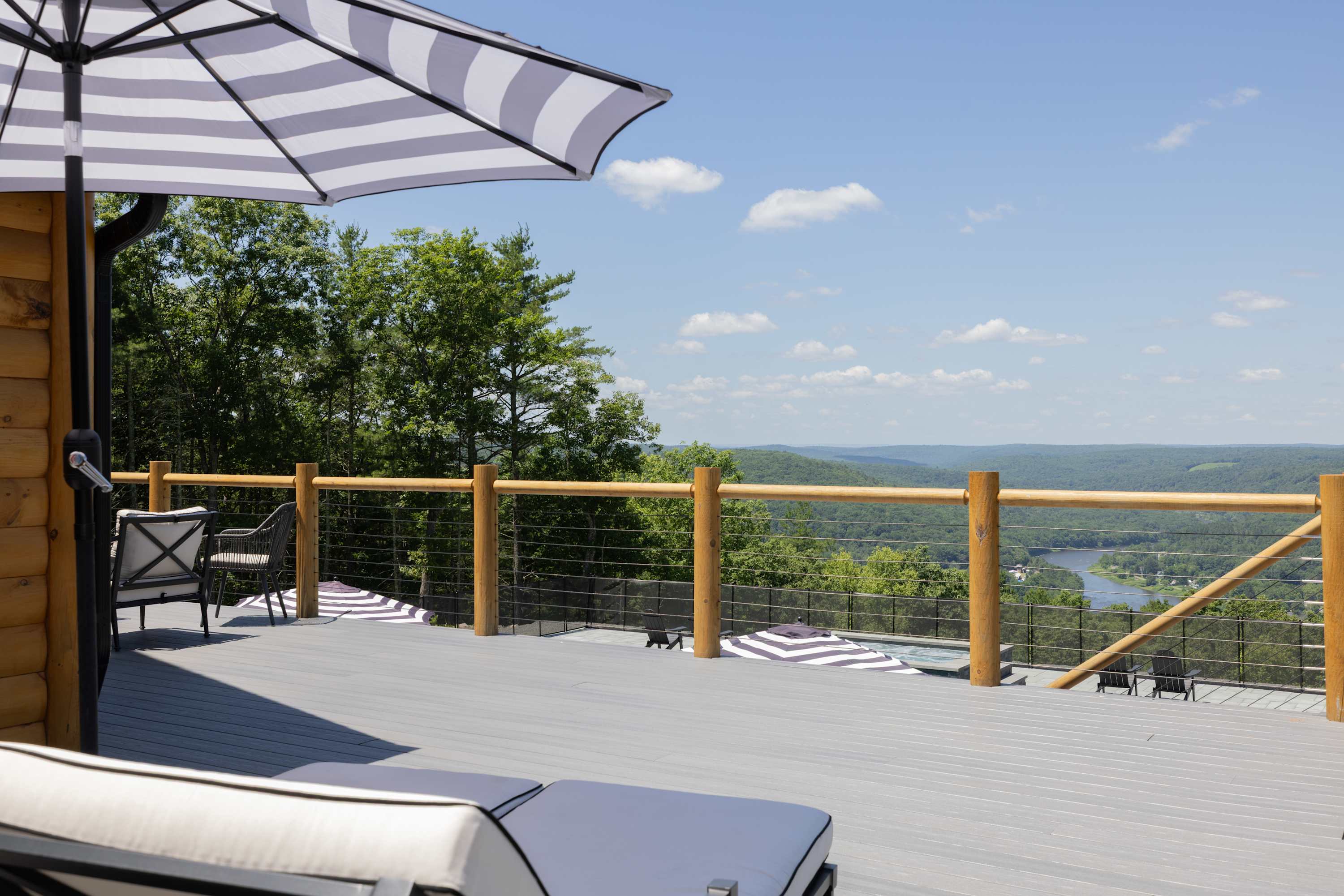 A wide gray composite deck with a wooden railing and cable infill provides breathtaking panoramic views of a vast green valley and a winding river in the distance. A striped umbrella and outdoor lounge chairs are visible.