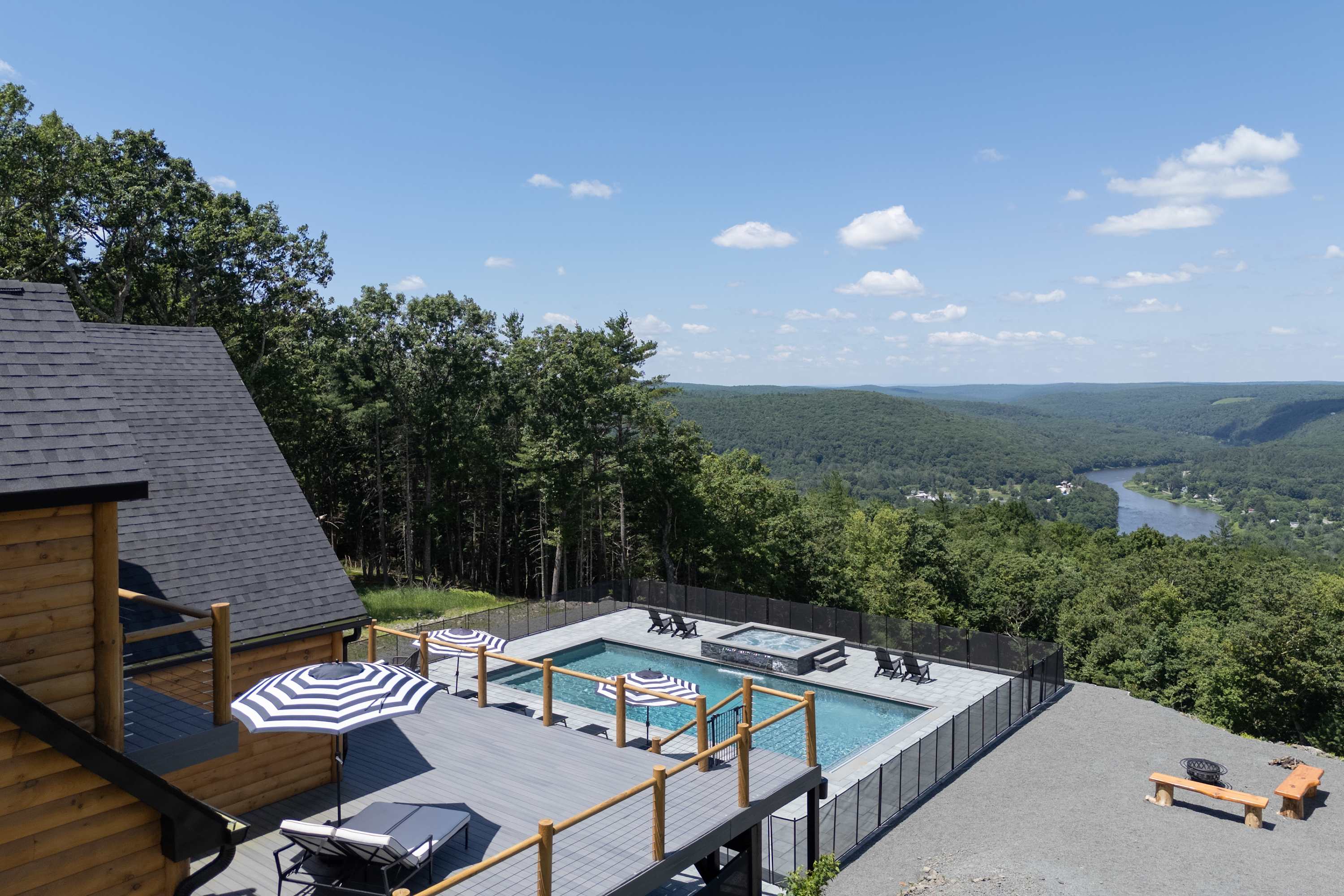 Panoramic aerial view showcasing a luxurious wooden lodge, a large pool and hot tub area, and a spacious deck, all set against a stunning backdrop of rolling green hills and a winding river.