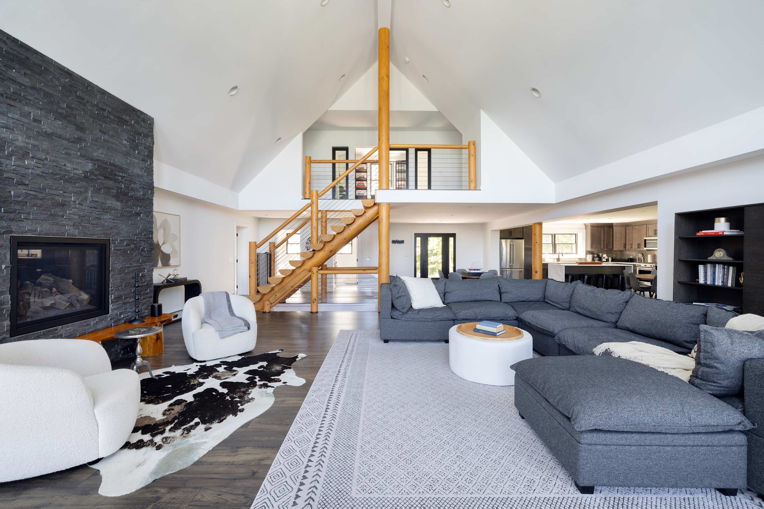 Open-concept living room with a vaulted ceiling, dark wood floors, and a prominent log-style wooden staircase leading to an upper level. A large grey sectional sofa and stone fireplace anchor the space.