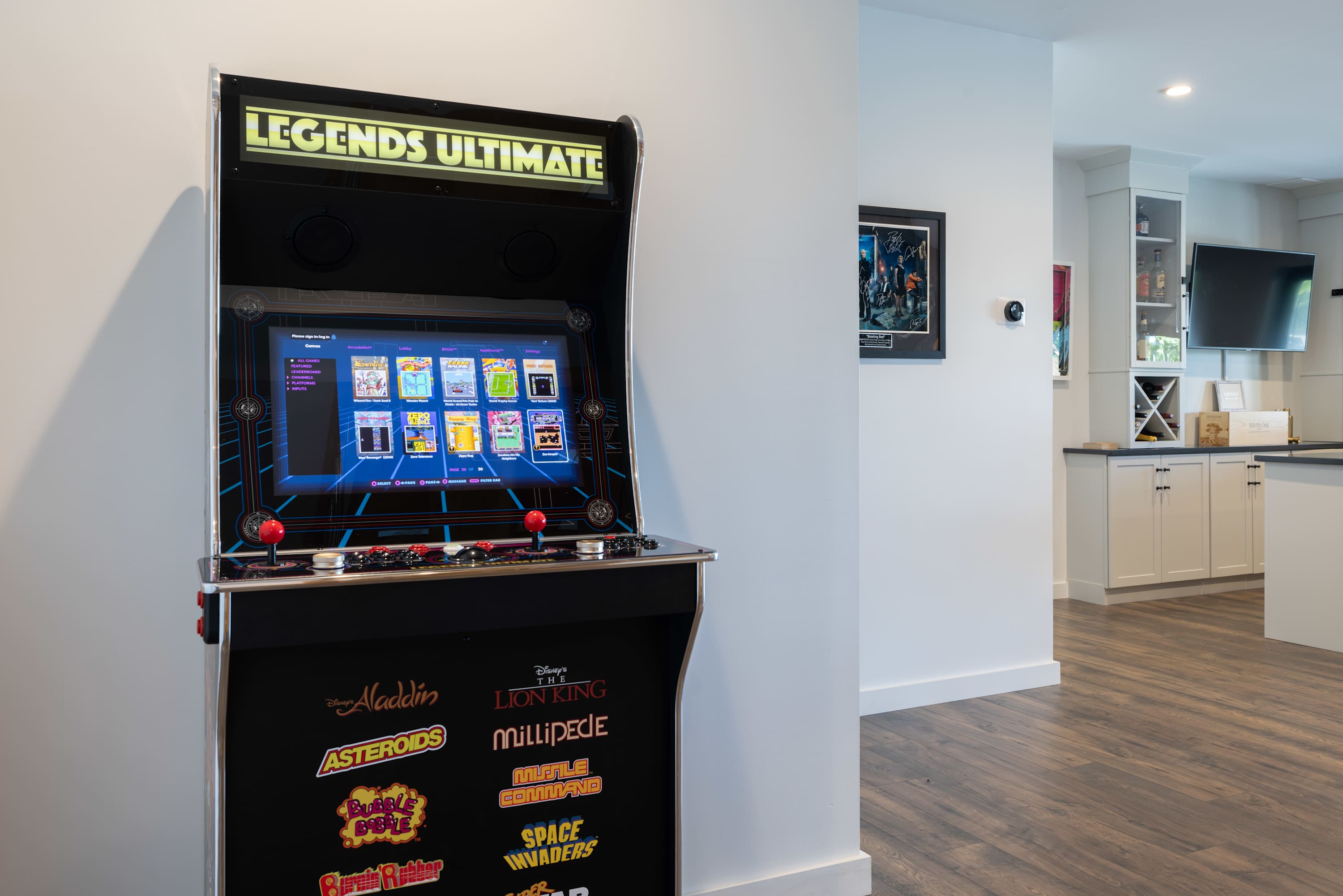 Vintage-style arcade game machine with "Legends Ultimate" branding, featuring a large screen displaying classic games, positioned in a bright white hallway next to a bar area.