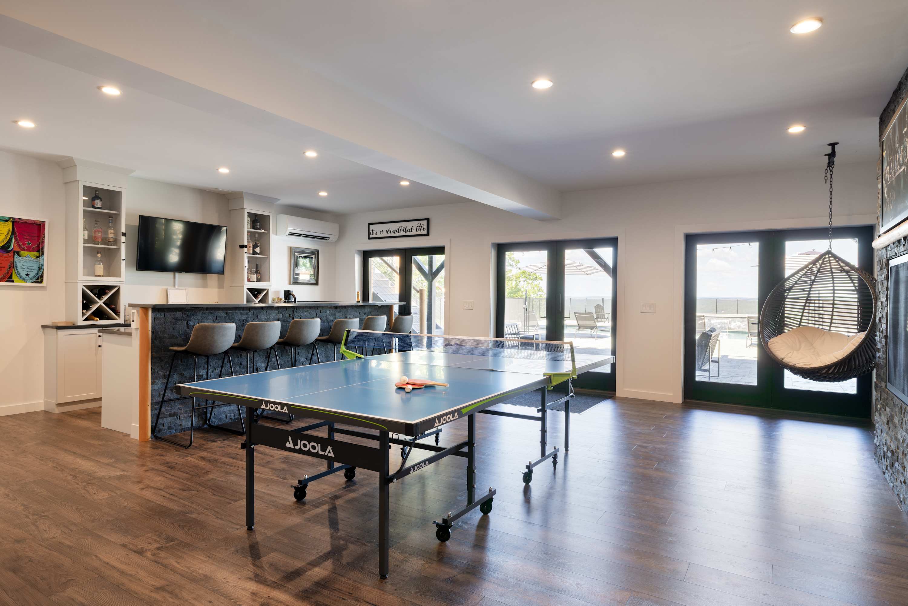 Expansive game room with dark wood floors, a ping pong table in the center, and a modern bar area with stools on the left. Large glass doors lead to an outdoor space, and a hanging chair adds a unique touch.