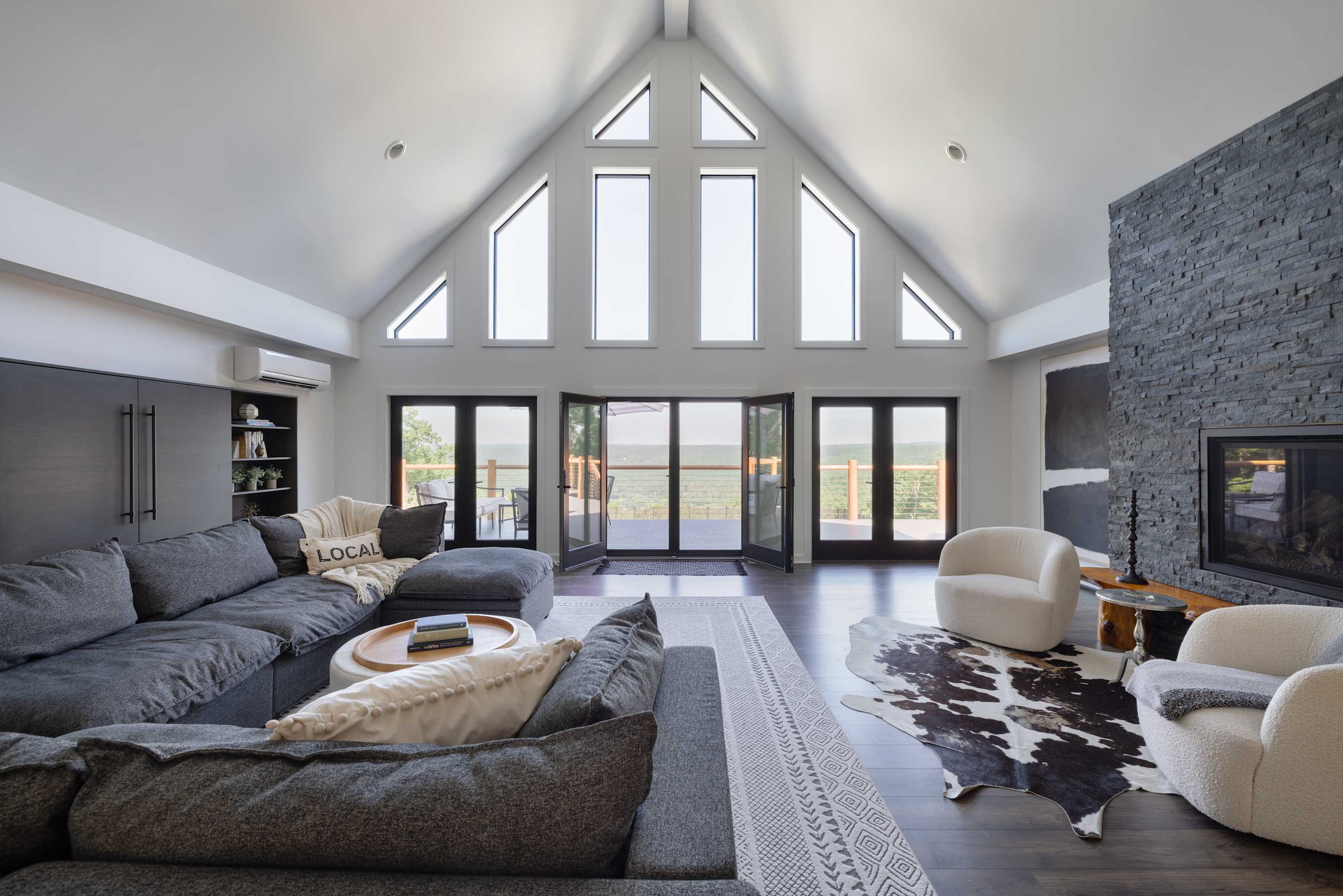 Spacious living room with a vaulted ceiling, large triangular windows, and dark wood floors. A large grey sectional sofa faces a modern stone fireplace, complemented by a cowhide rug and white armchairs.