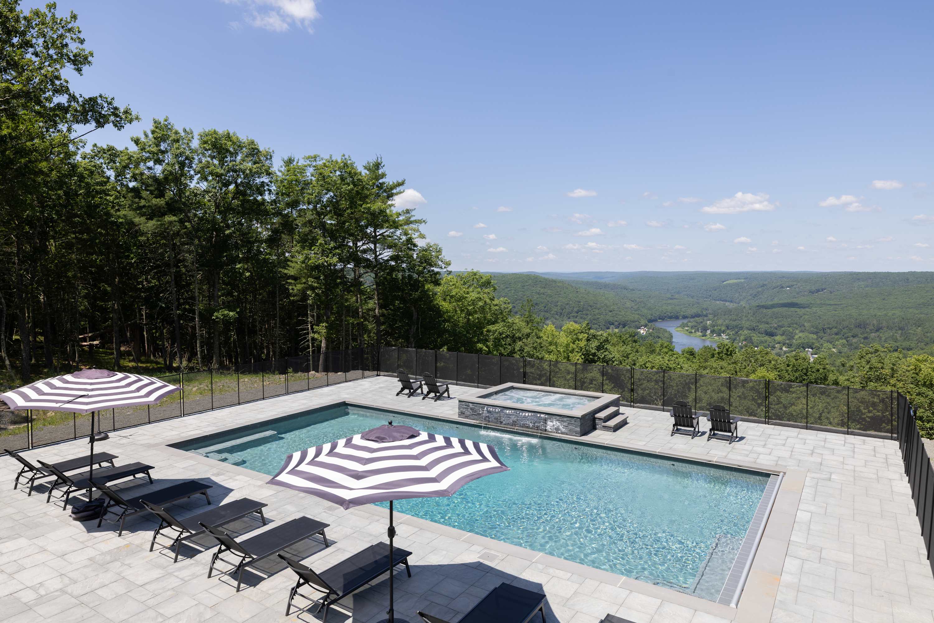 A large rectangular swimming pool and an elevated hot tub on a gray paver patio, furnished with black lounge chairs and striped umbrellas. The area is enclosed by a black fence, offering expansive views of a lush green valley and a distant river.