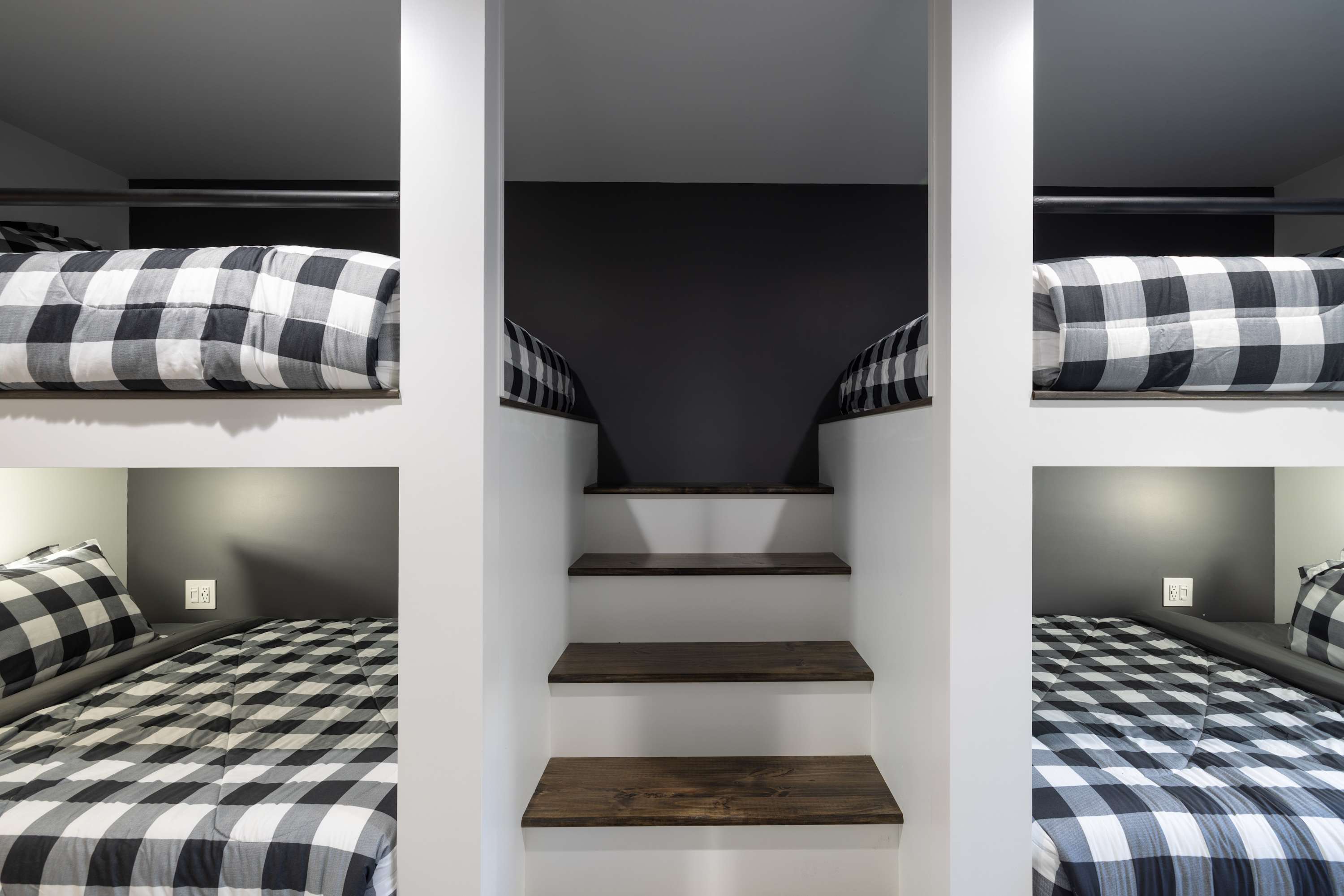 Close-up view of the built-in bunk beds with black and white buffalo check bedding, featuring wooden steps leading to the upper bunks. The walls are white with a dark accent wall at the top of the stairs.