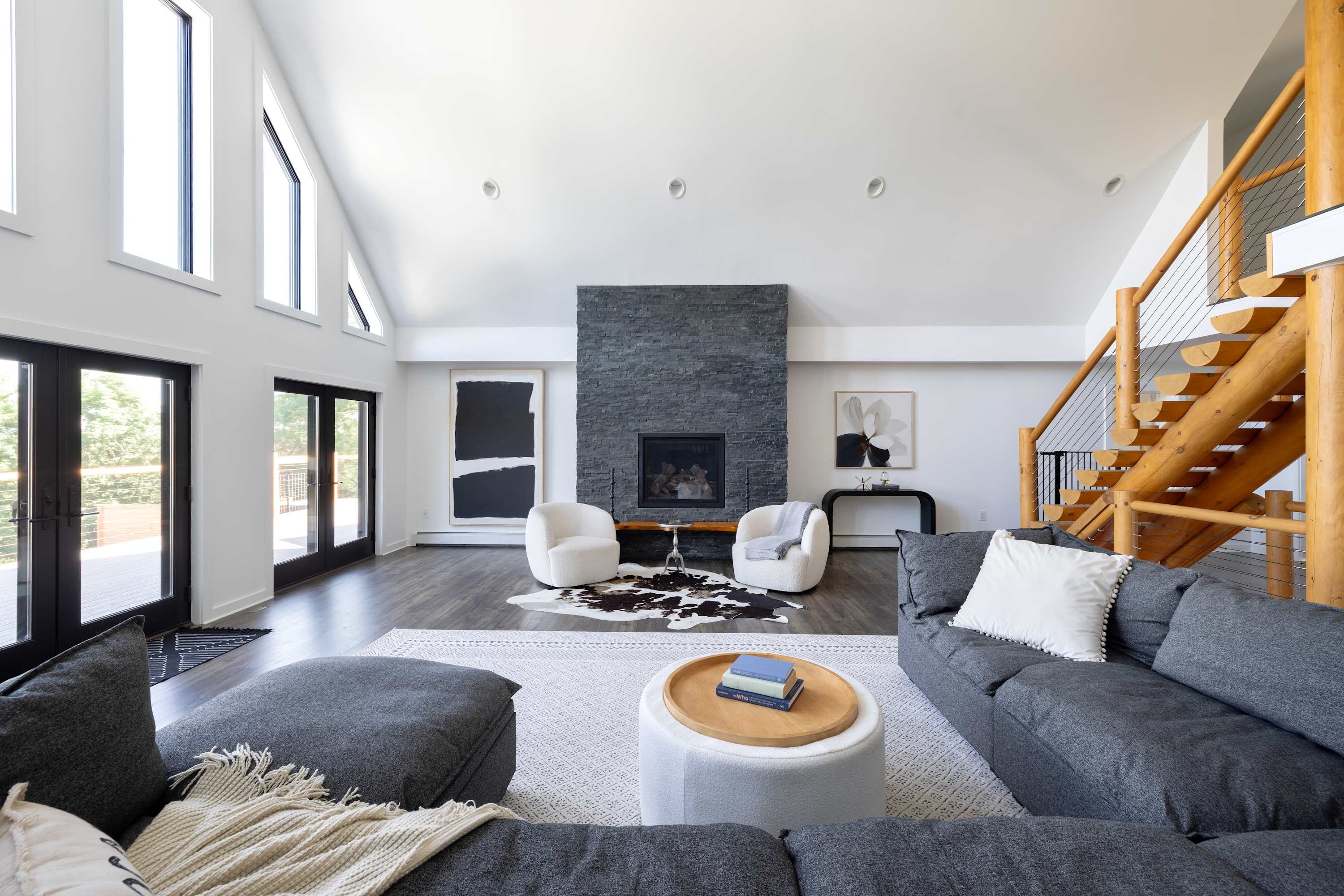Spacious living room with a high vaulted ceiling and large windows, featuring a large grey sectional sofa, a modern stone fireplace, and a cowhide rug, creating a cozy yet grand atmosphere.