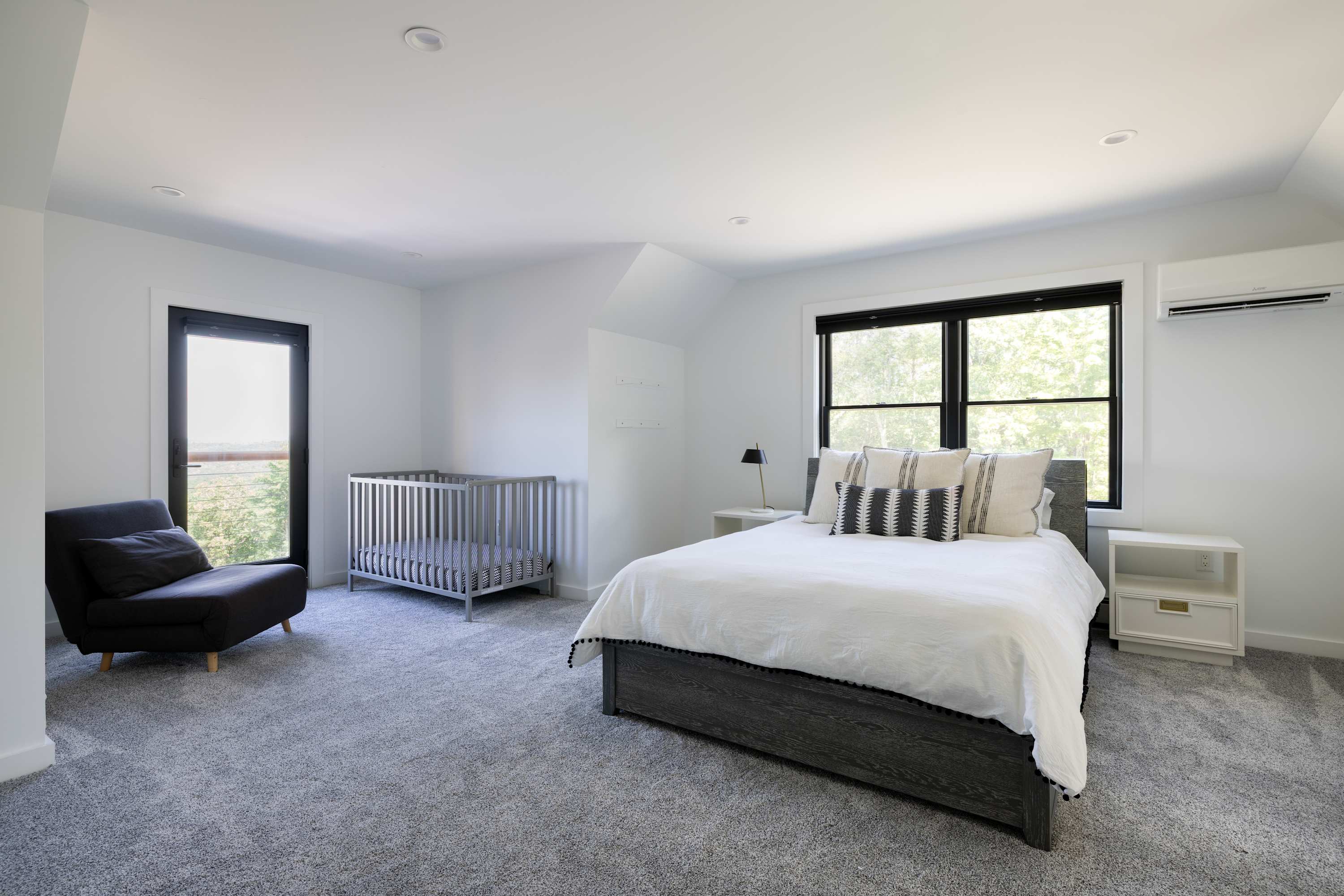 Bright bedroom with light gray carpet, a dark gray bed frame with white bedding, and a matching crib. A dark armchair sits near a black-framed glass door leading to a balcony.