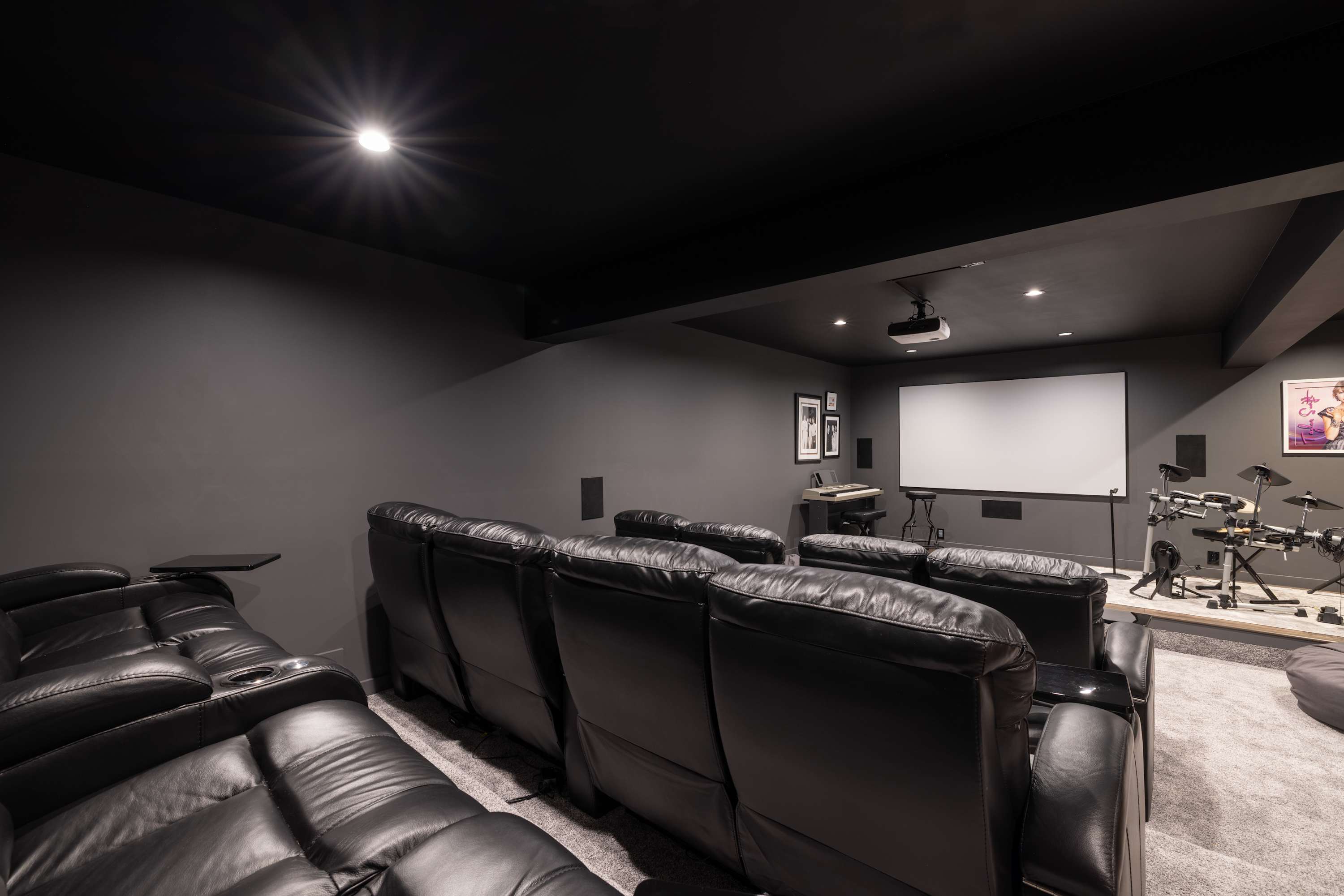 Dark-themed home theater with multiple rows of black leather reclining seats, a large projection screen, and recessed lighting, featuring musical instruments like a drum set and keyboard in the corner.