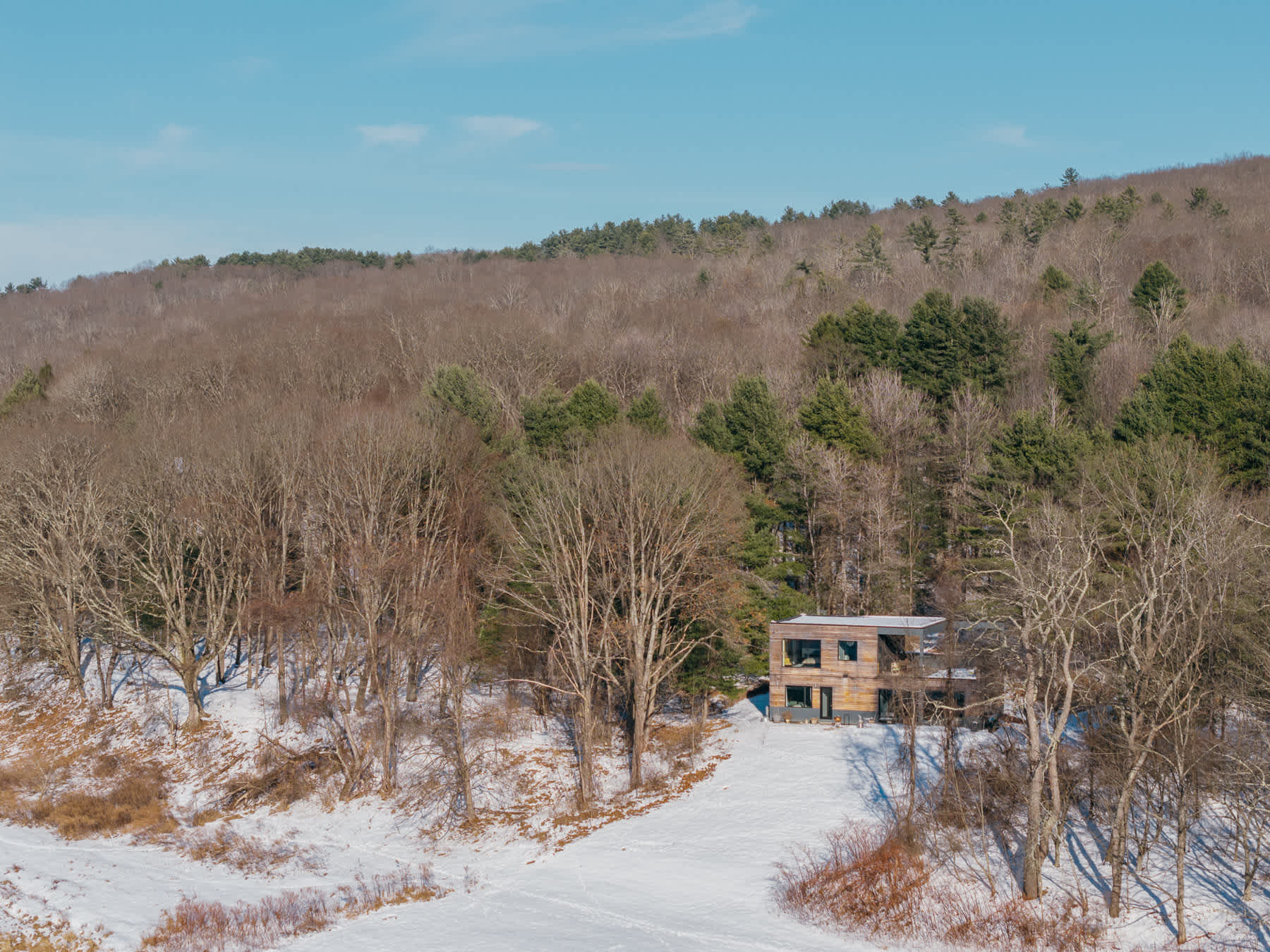 Aerial view of a contemporary wooden-sided house situated on a snowy bank, surrounded by a mix of bare and evergreen trees, with a wide, partially frozen river in the foreground and forested hills beyond.