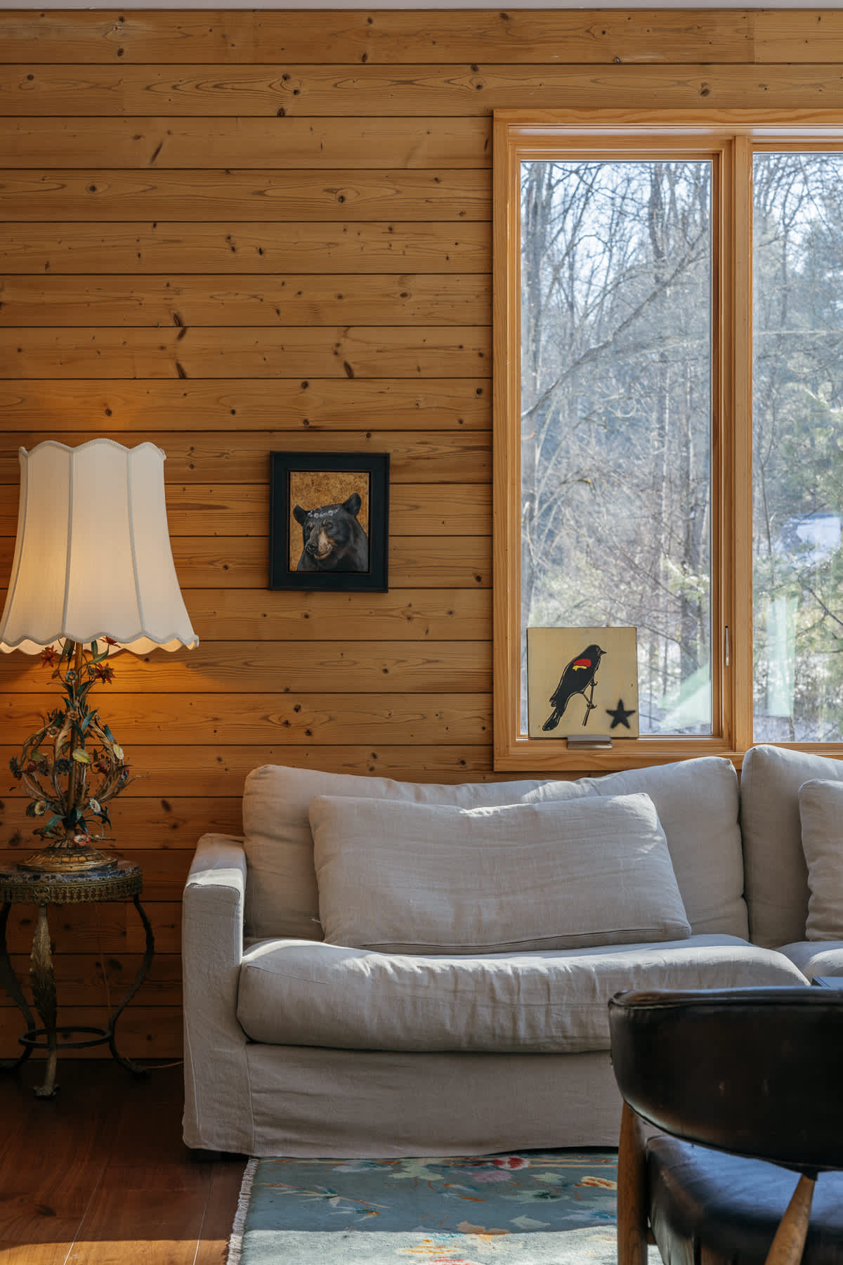 A corner of a living room featuring light wood paneling, a comfortable light-colored sofa, a vintage-style lamp, and a window looking out to a snowy, wooded scene.