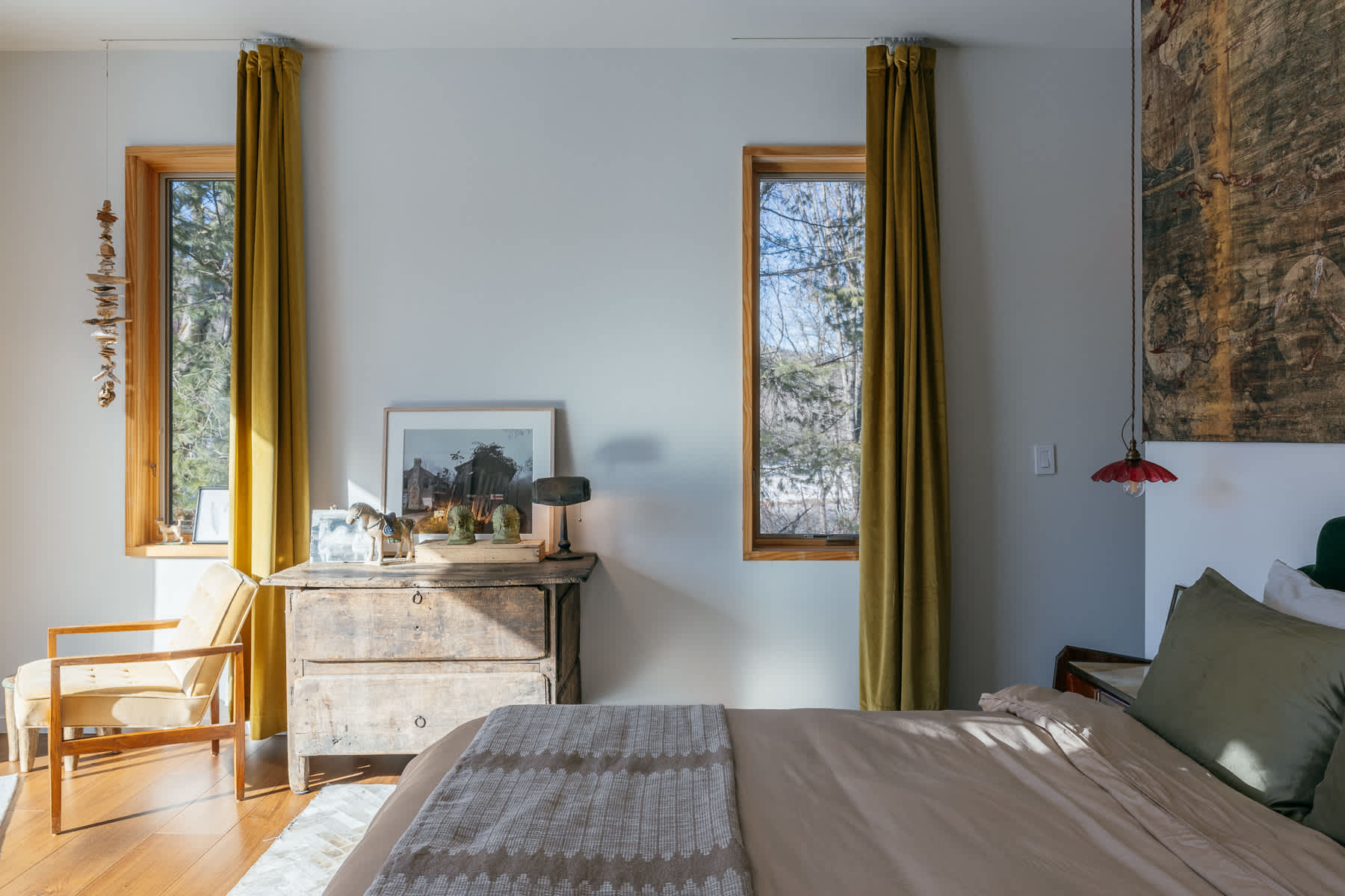 Bright bedroom with light gray walls, two tall windows framed by mustard yellow curtains, a rustic wooden dresser, and a bed with a patterned throw, offering a view of the outdoors.