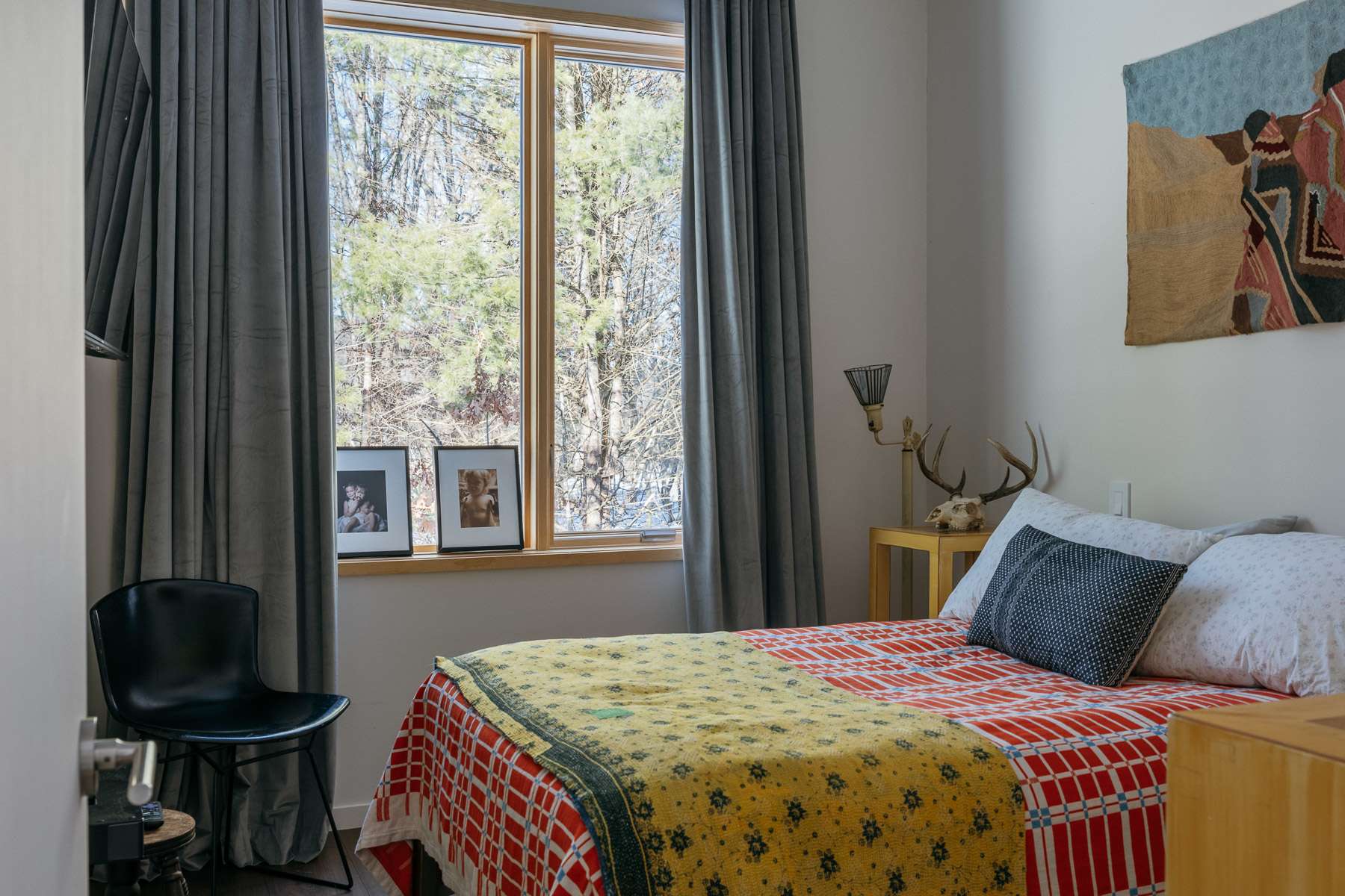 A bright bedroom with white walls, featuring a bed adorned with a colorful patterned quilt. A window with grey curtains overlooks a snowy landscape, and a tapestry hangs above the bed.