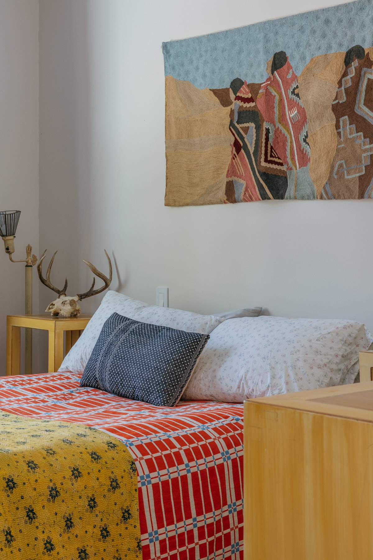 A bedroom featuring a bed with a vibrant, patterned quilt and a large tapestry hanging above. A side table holds decorative antlers and a skull, with a window visible in the background.