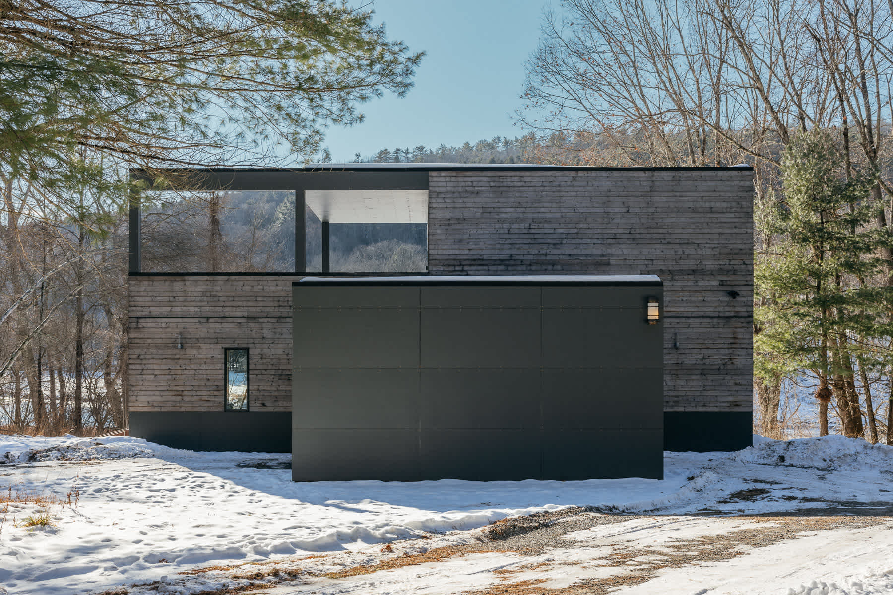Contemporary house with horizontal wood siding and a prominent dark gray panel structure, featuring a large horizontal window offering views of the snowy, wooded landscape.