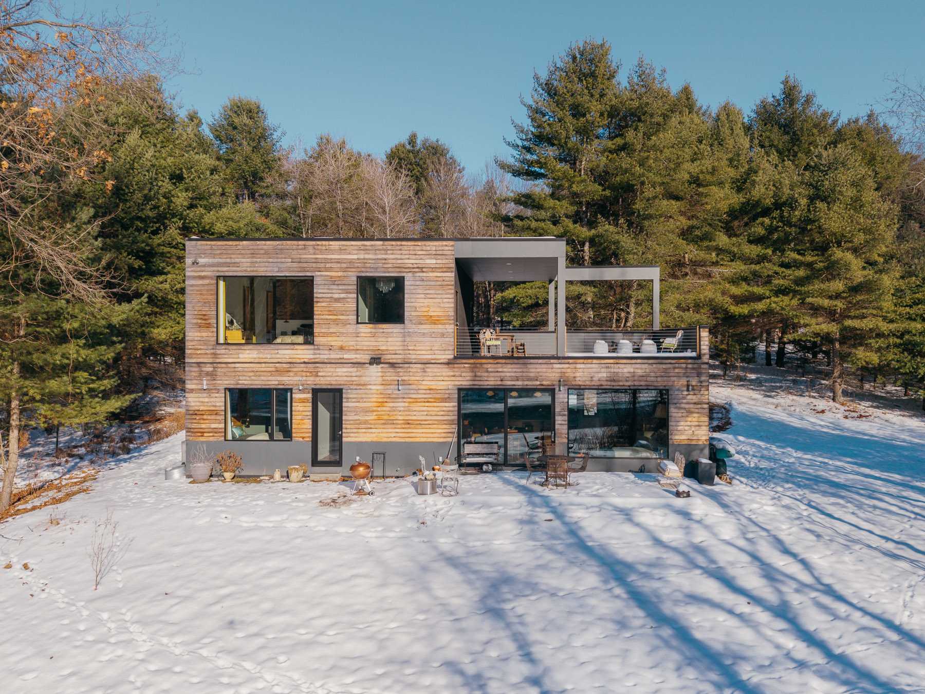 Aerial view of the modern house's rear facade, showcasing its wood siding, large windows, and expansive deck with outdoor furniture, overlooking a snowy landscape and a river.