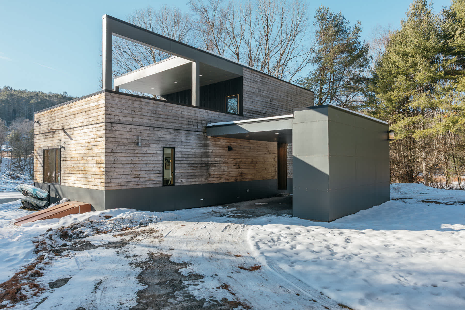 Contemporary house with horizontal wood siding and dark gray accents, featuring a covered entrance and a second-story balcony, surrounded by snow and winter trees.