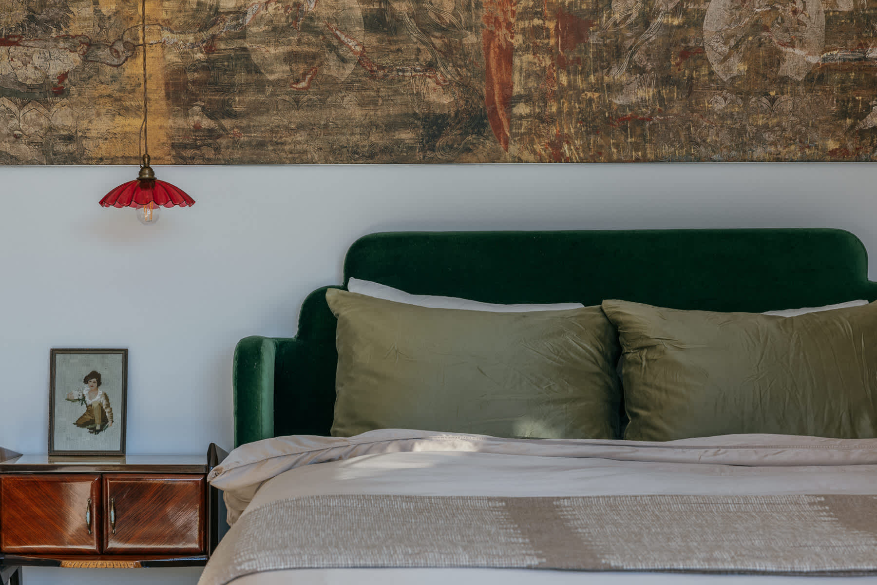 Close-up of a luxurious bedroom with a dark green velvet headboard, olive green pillows, and a light-colored duvet, accented by a small red pendant light and a vintage nightstand.