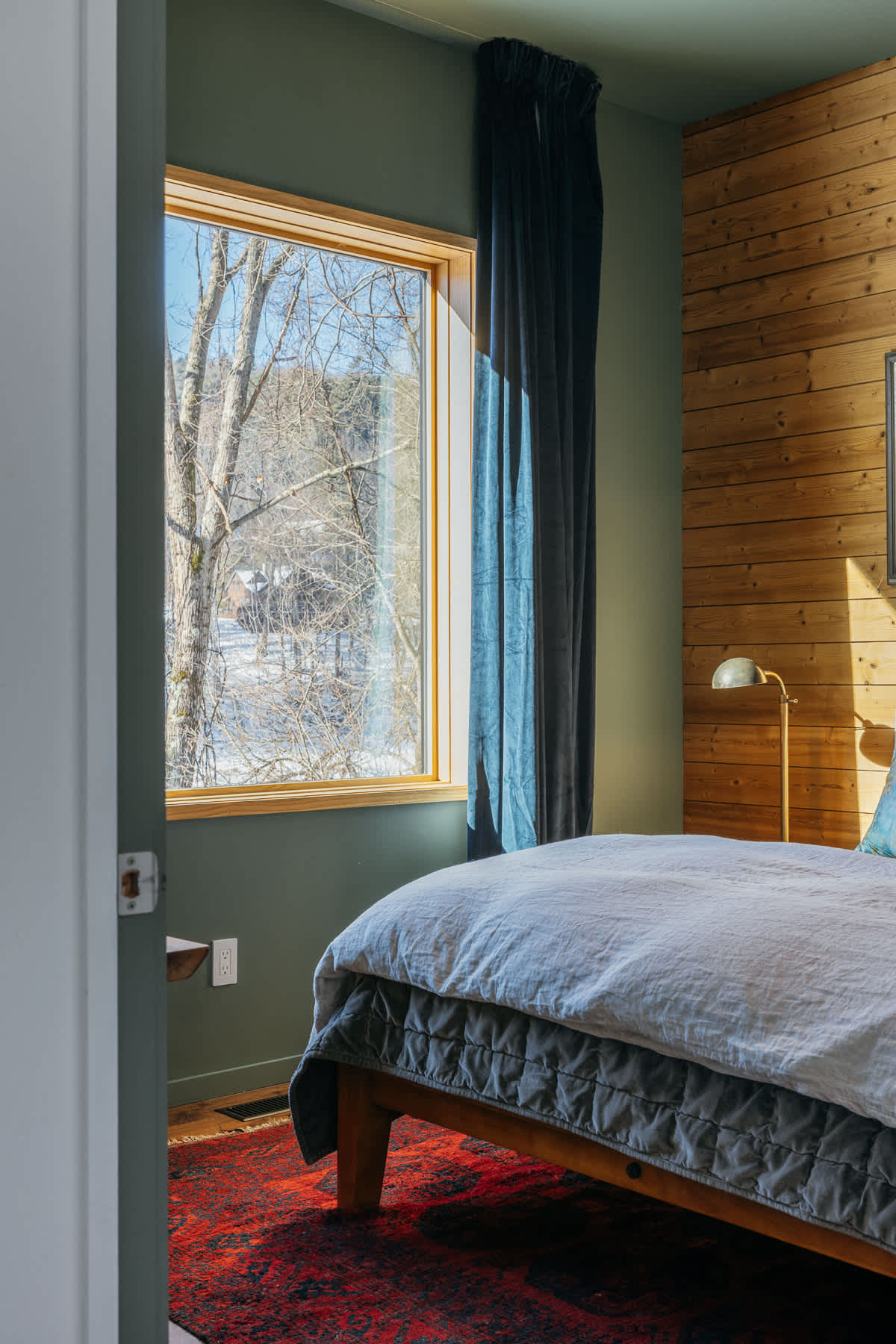 A view into a bedroom with dark green walls and a large window framed by dark blue curtains, revealing a snowy outdoor landscape. A bed with a white duvet and a red patterned rug are visible.