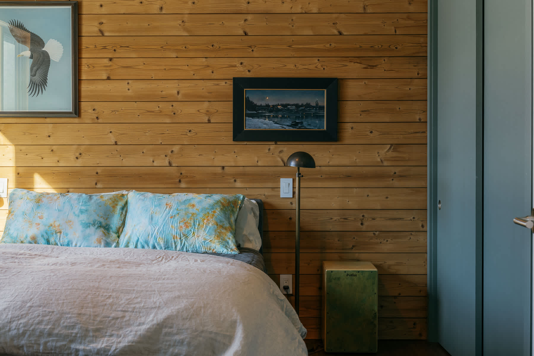 A bedroom with a bed positioned against a warm wooden plank accent wall, featuring two framed artworks. A modern floor lamp and a small side table are next to the bed.