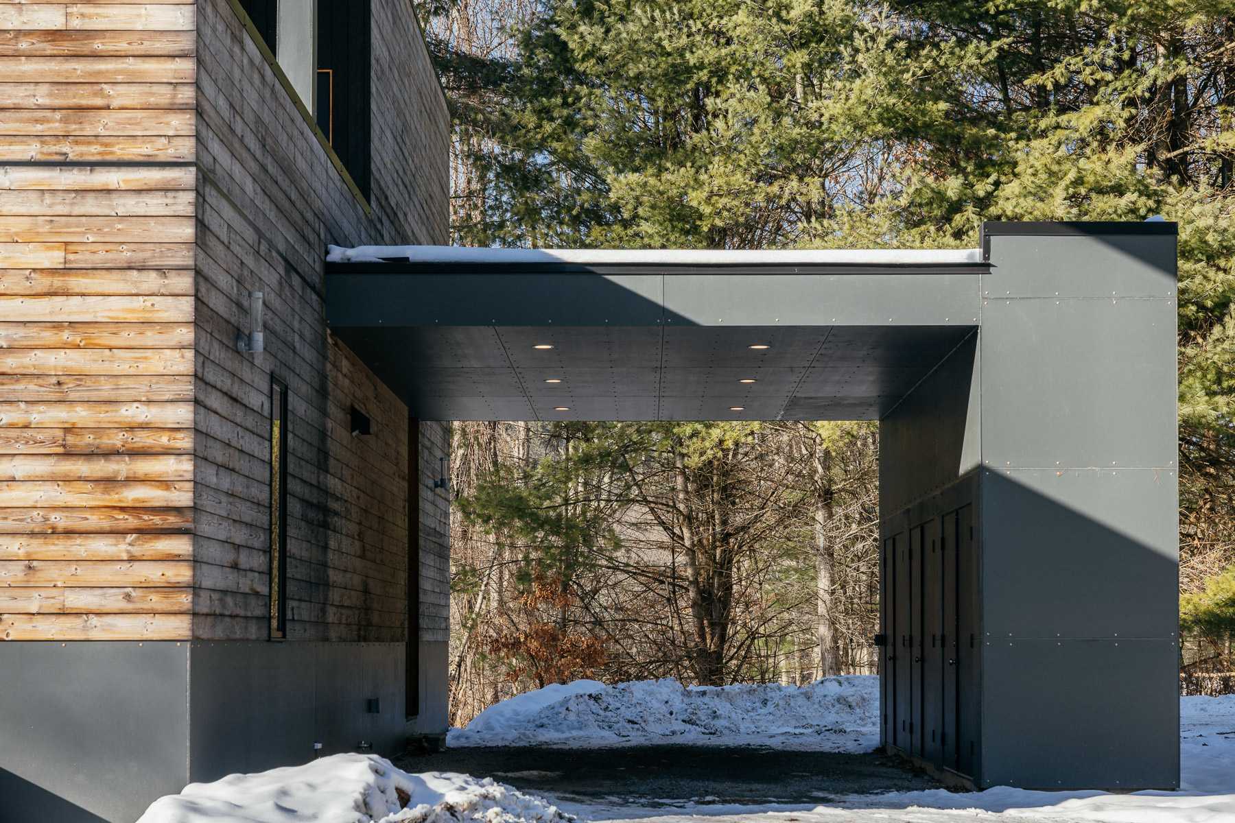 A modern covered walkway or carport with dark grey paneling and recessed lighting, connecting to a building with horizontal wooden siding. Snow is visible on the ground.