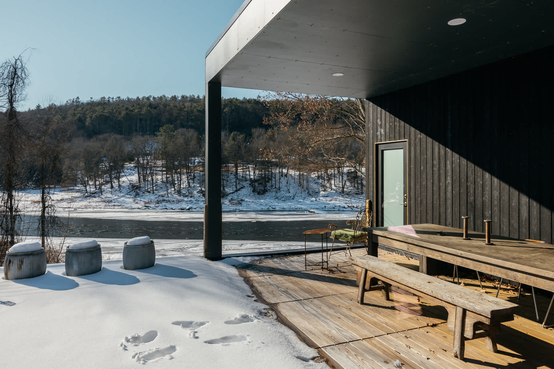 Ground-level wooden deck with a picnic table and benches, adjacent to a dark-sided wall, offering views of a snowy riverbank and partially frozen river.