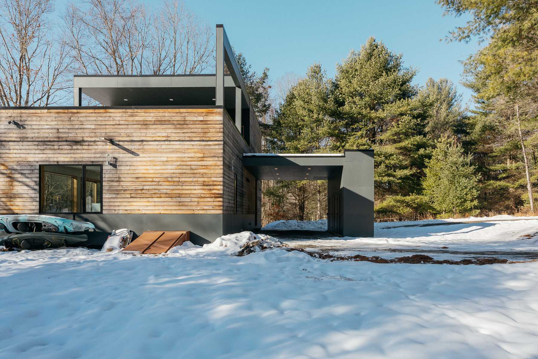 Modern house with horizontal wood siding and dark gray panels, featuring a covered carport entrance, set in a snowy landscape with bare trees and evergreens in the background.