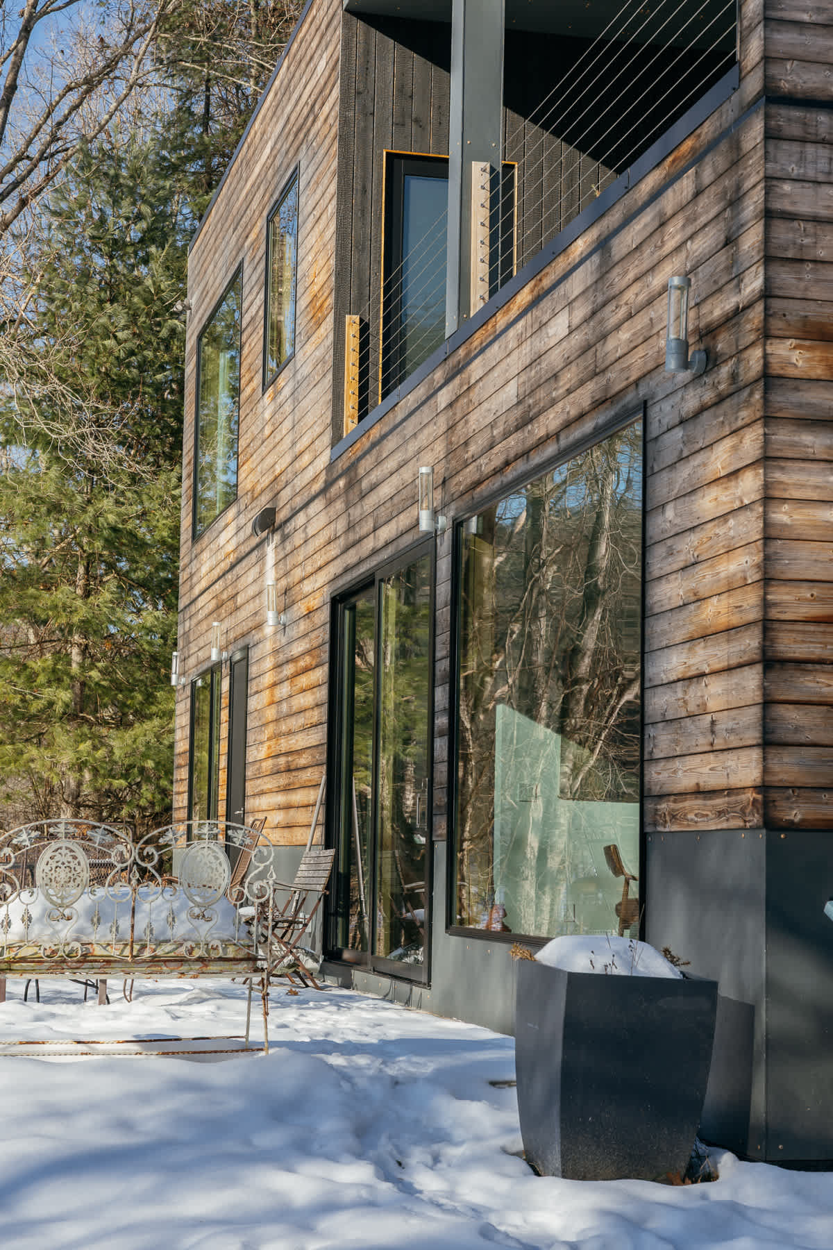 The side of a modern house with horizontal wooden siding and large black-framed windows, overlooking a snow-covered landscape. An ornate white metal bench sits on the snowy ground in the foreground.
