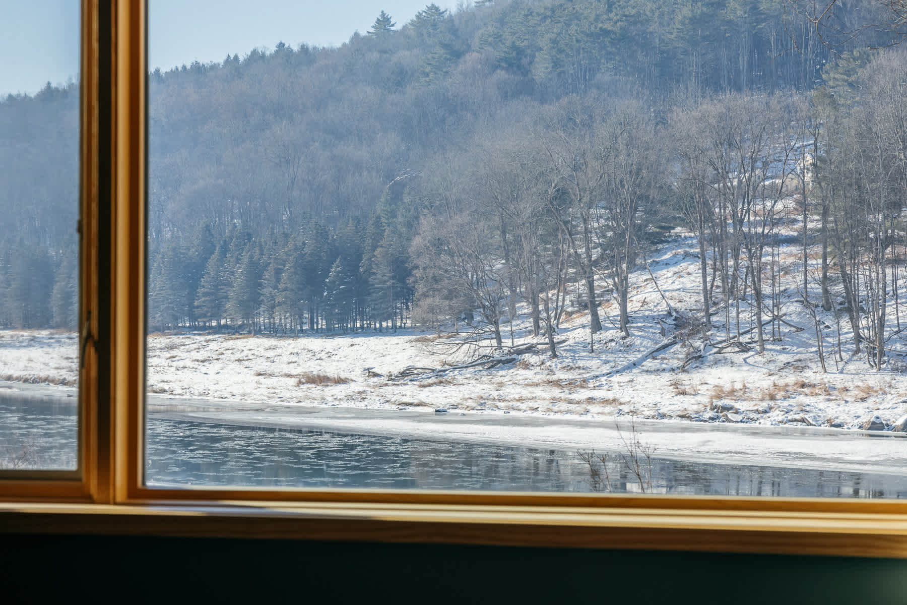 A close-up view through a window showcasing a serene winter landscape with a partially frozen river, snow-dusted banks, and a dense forest on the distant hillside under a clear sky.