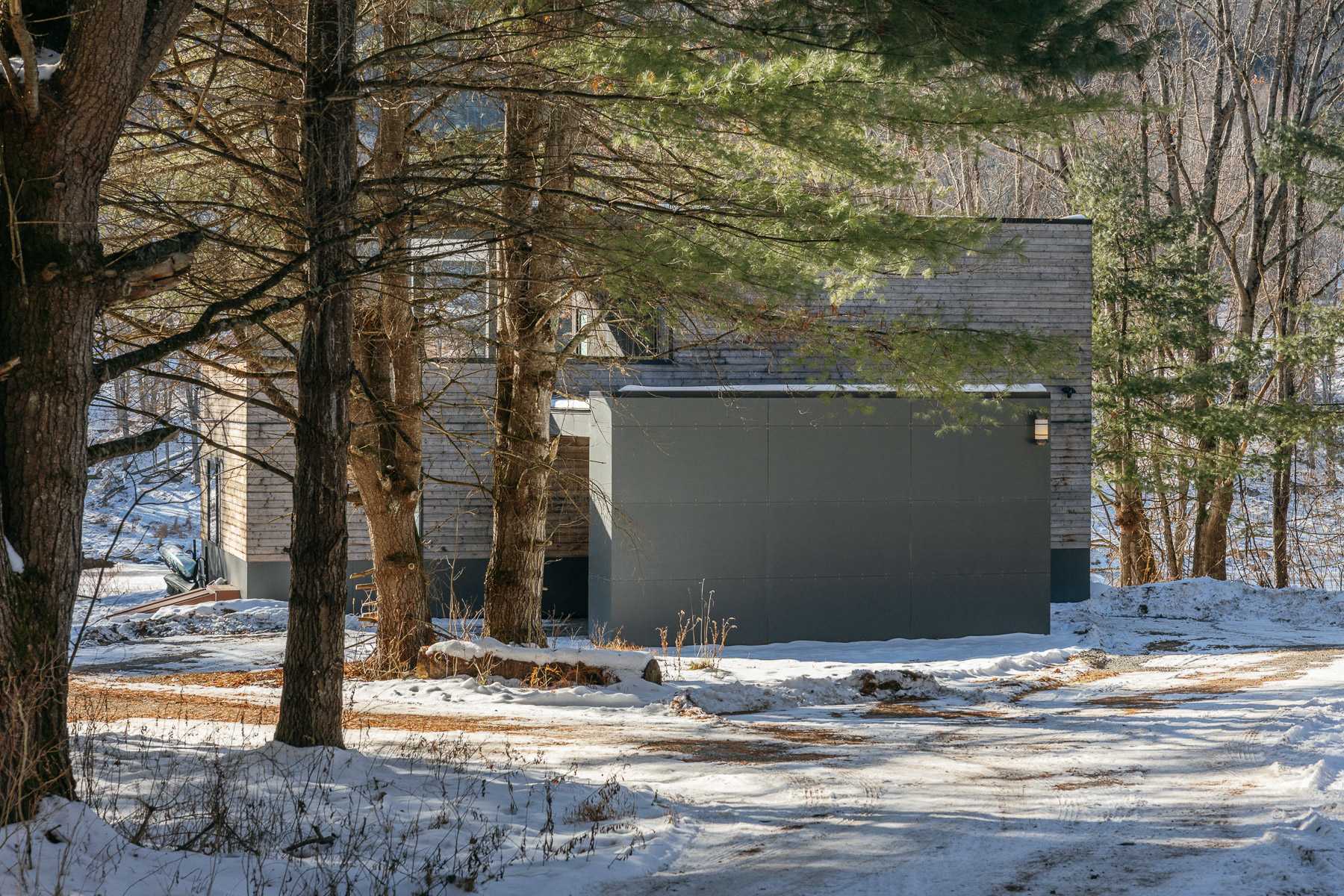 The modern house, featuring wooden siding and dark grey paneling, is partially hidden among bare trees in a snow-covered setting.